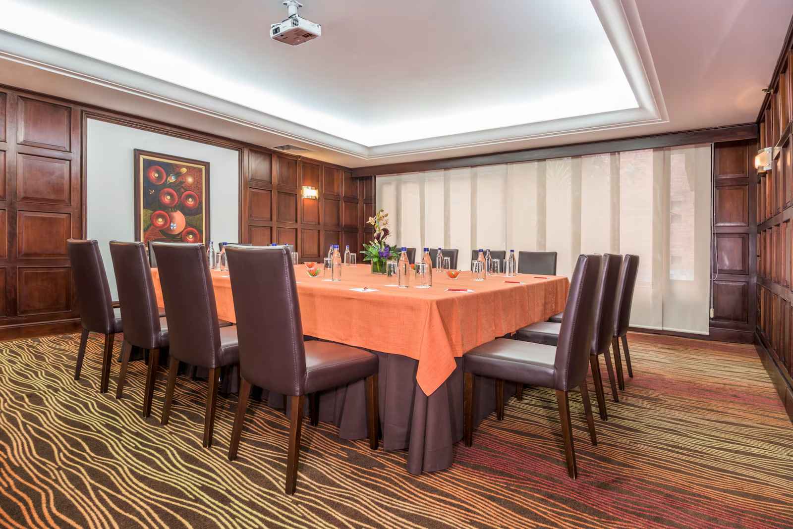  Salón comedor con una amplia mesa de madera, cubierta con un mantel naranja y rodeada de sillas de cuero. Las paredes están cubiertas con madera. Hay un gran ventanal cubierto con una persiana blanca.