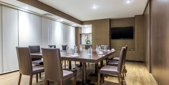  Salón de reunión con una amplia mesa de madera, y rodeada de sillas de cuero. Las paredes están cubiertas con madera. Hay un gran ventanal cubierto con una persiana blanca, y un televisor colgado en una pared.