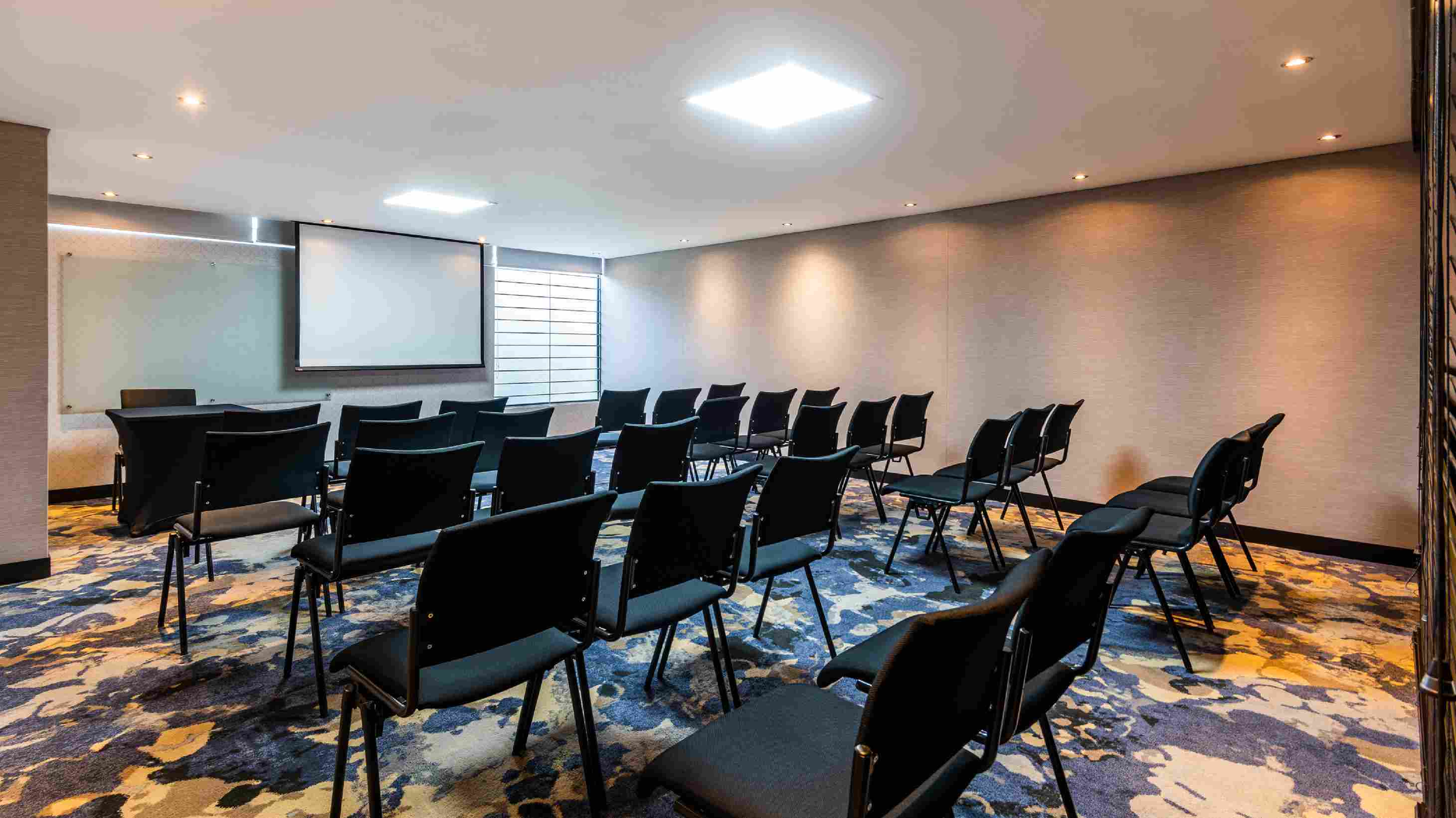 Salón pequeño con por lo menos dos docenas de sillas metálicas, todas de frente a una pantalla de videobeam.