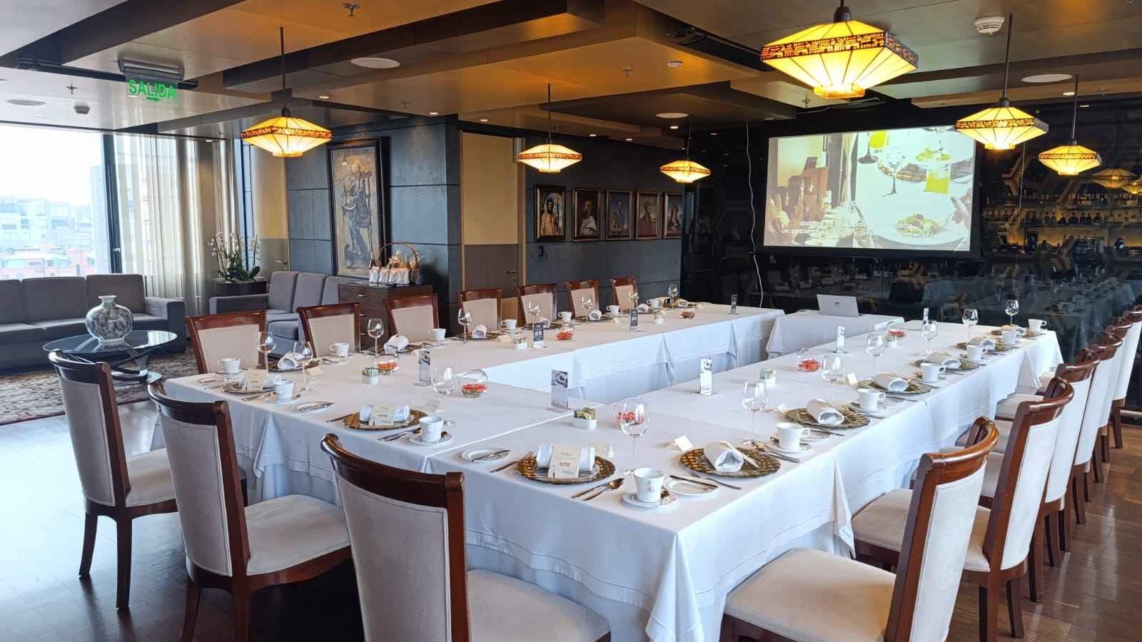  Salon con una mesa en forma de U cubierta con mantel blanco y con copas de cristal y platos sobre ella. Hay una pantalla gigante al fondo.