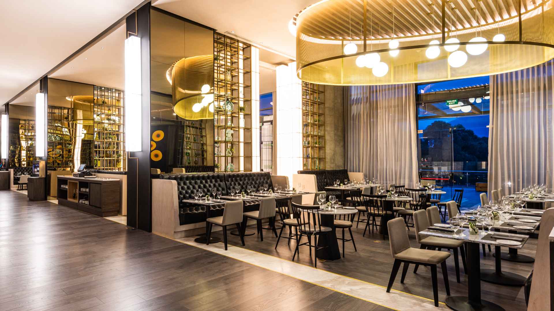 Salón elegante iluminado con lámparas en el techo, y en el que hay varias mesas cuadradas rodeadas por poltronas y sillas. Hay varias mesas redondas rodeadas por butacos. Hay un ventanal hacia la calle.