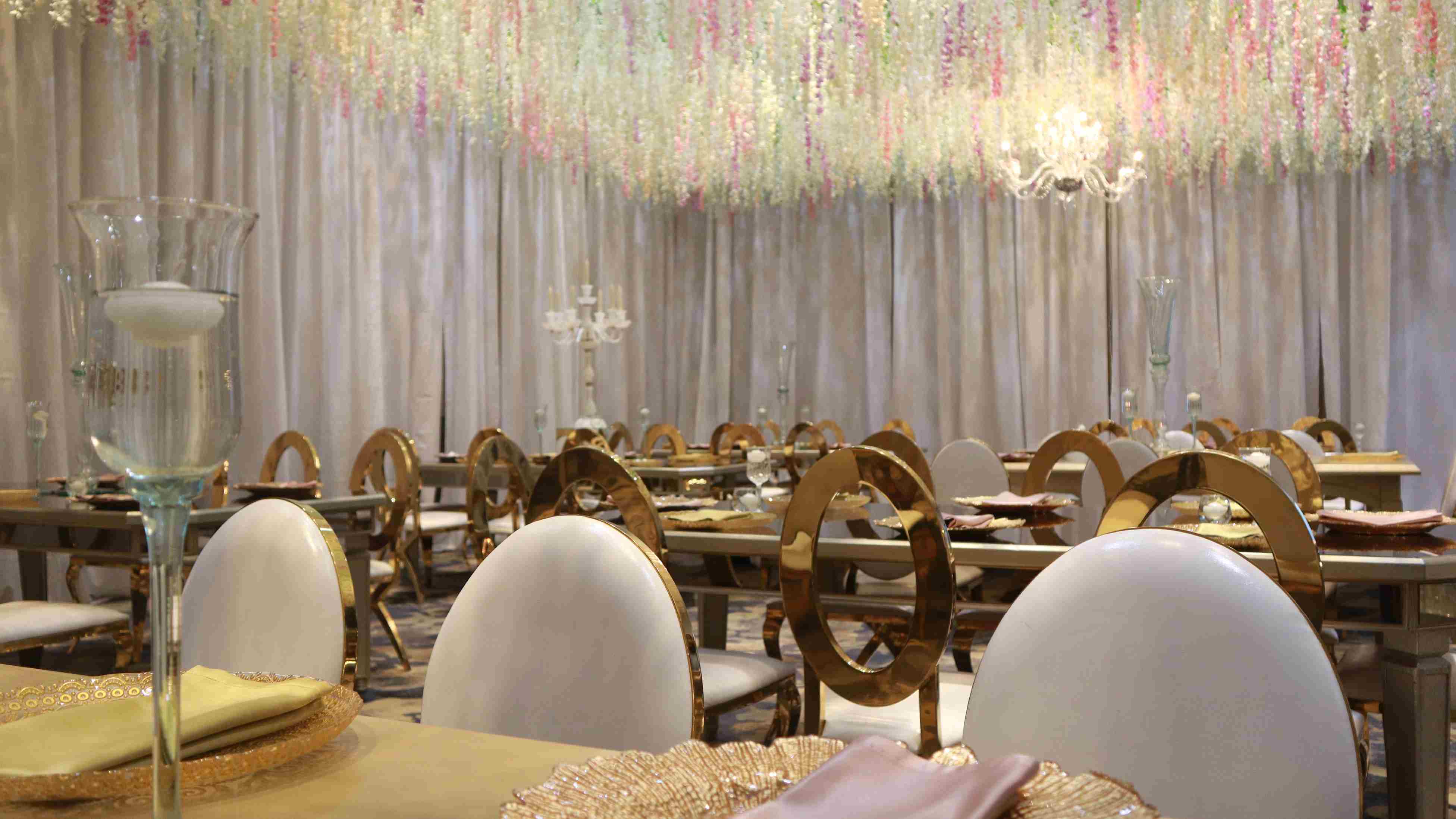 Close-up of an elegantly decorated hall with tables and chairs featuring golden details, a chandelier, and a decorative ornament, such as a tall glass goblet, on each table. The curtains have a decorative detail made with shiny thread.