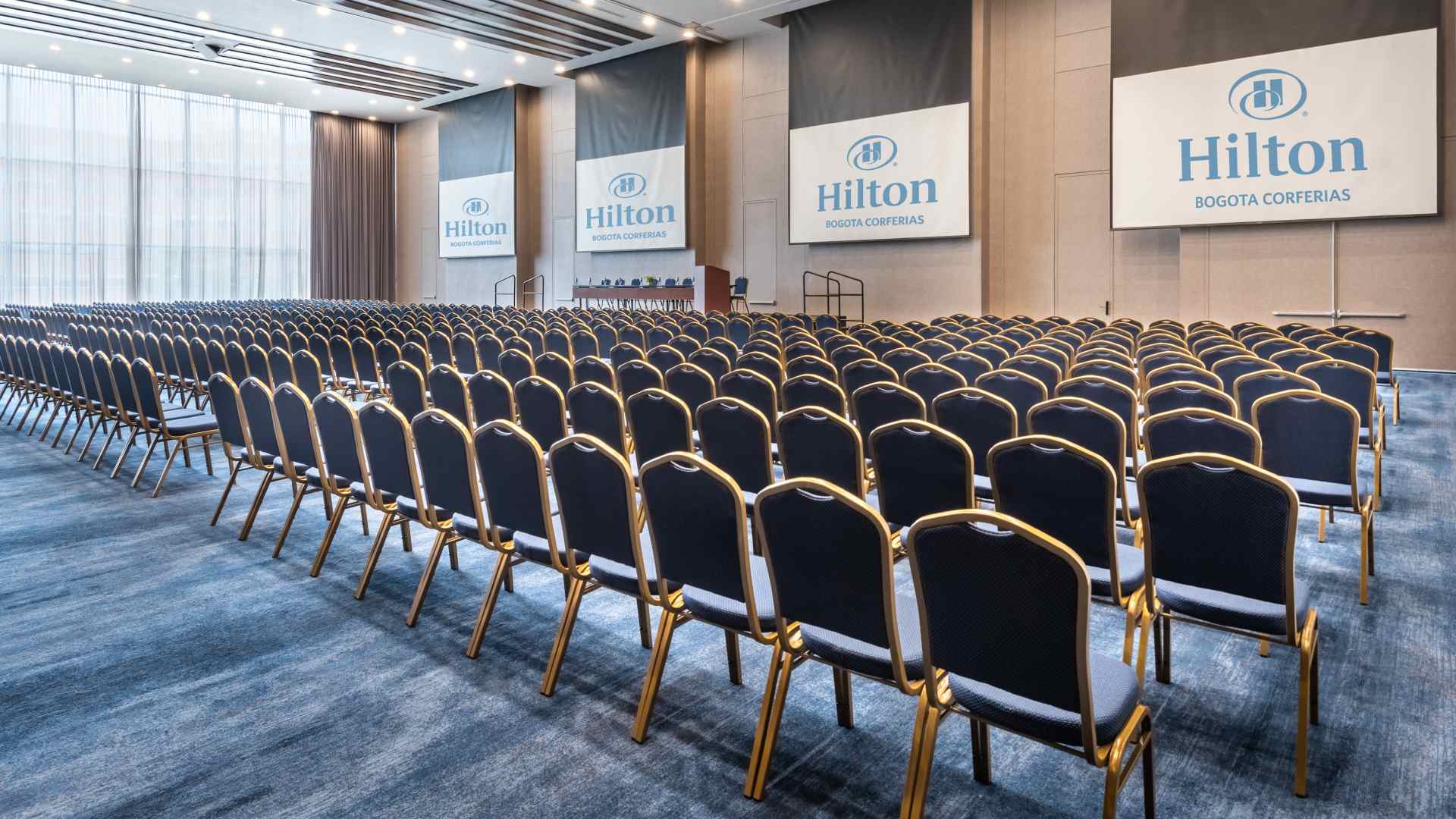 Gran salón con más de un centenar de sillas dispuestas freente a una pequeña tarima en la que hay una mesa con varias sillas. Destacan 4 pantallas de videobeam con leyendas que dicen: "Hilton Bogotá Corferias".