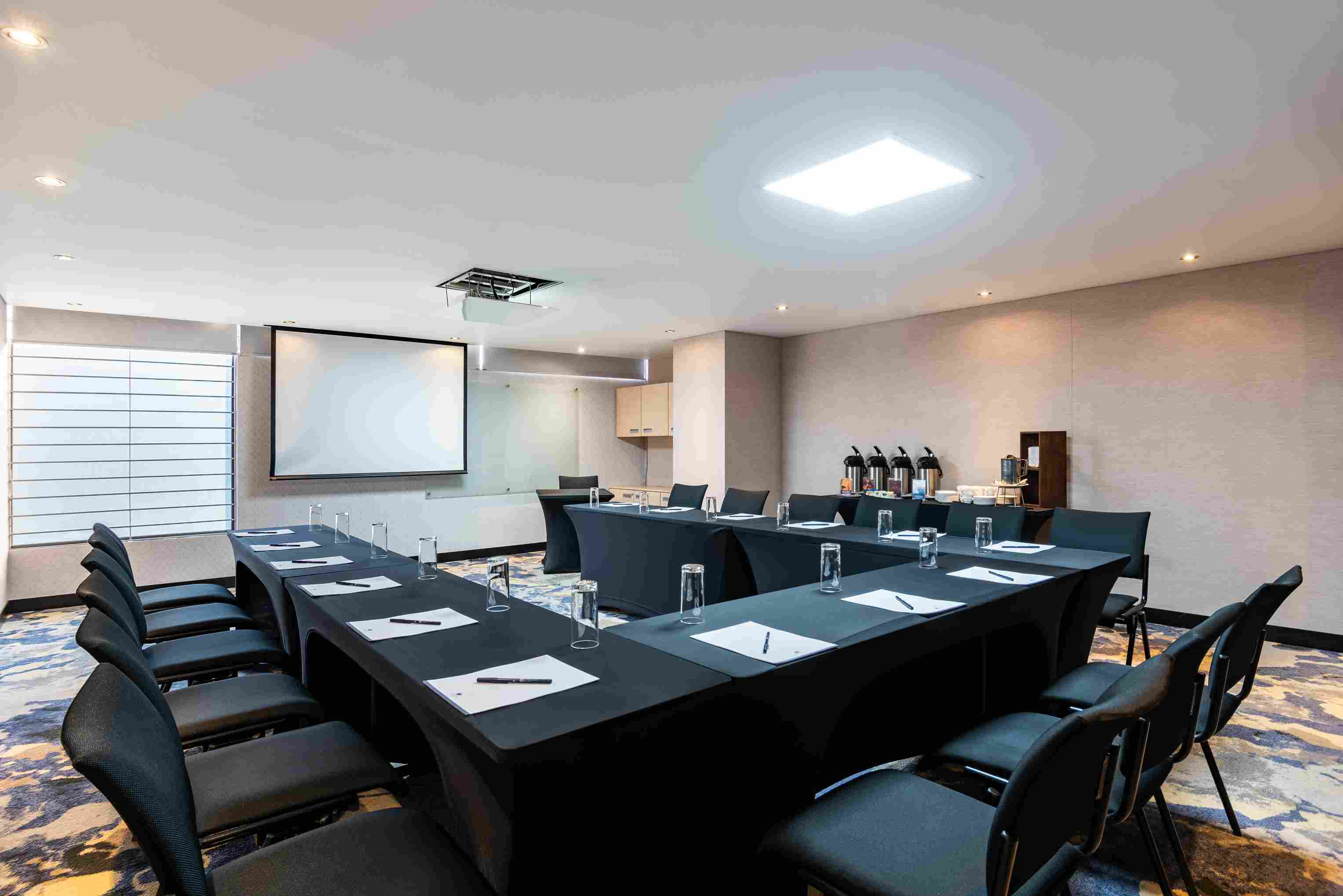 Salón pequeño con una mesa en U, rodeada por sillas. En la mesa hay vasos de cristal, esferos y papel. A un lado hay mesa con cafeteras y al fondo hay una pantalla de videobeam.