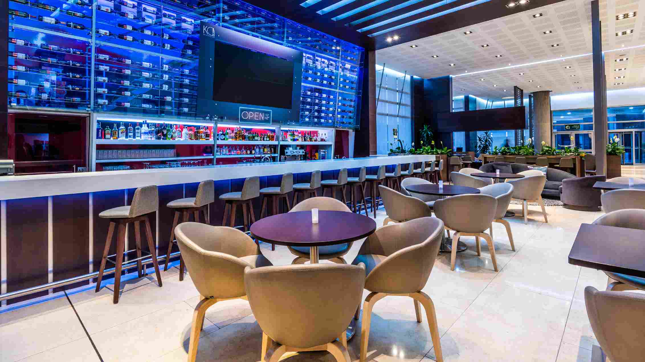  Large bar-style room featuring a bar, a glass wine cellar, and several round wooden tables with chairs around them.