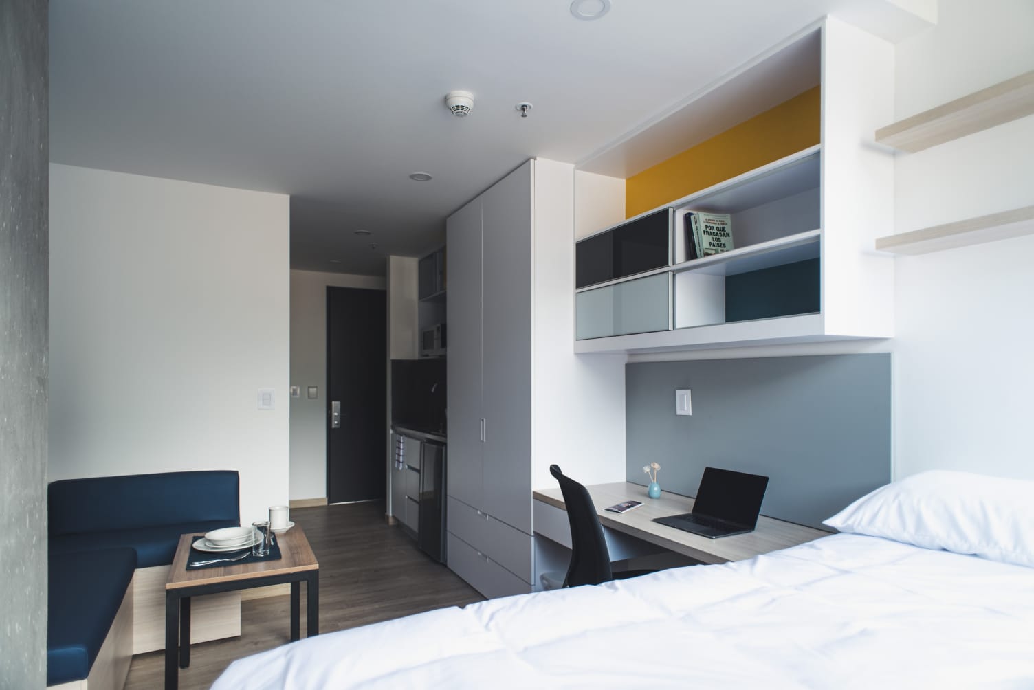  Habitación en la que hay una cama, una mesa de escritorio con un computador portatil, una mesa pequeña con platos y un vaso de cristal, y una especie de pequeña cocina.