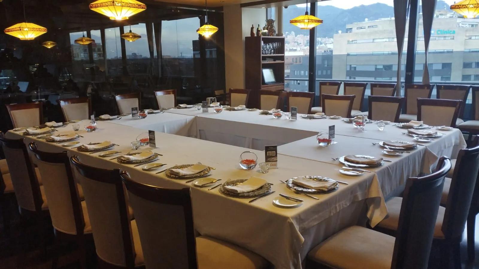 Salon con una mesa en forma de O cubierta con mantel blanco y con copas de cristal y platos sobre ella. Se ve un gran ventanal de fondo.