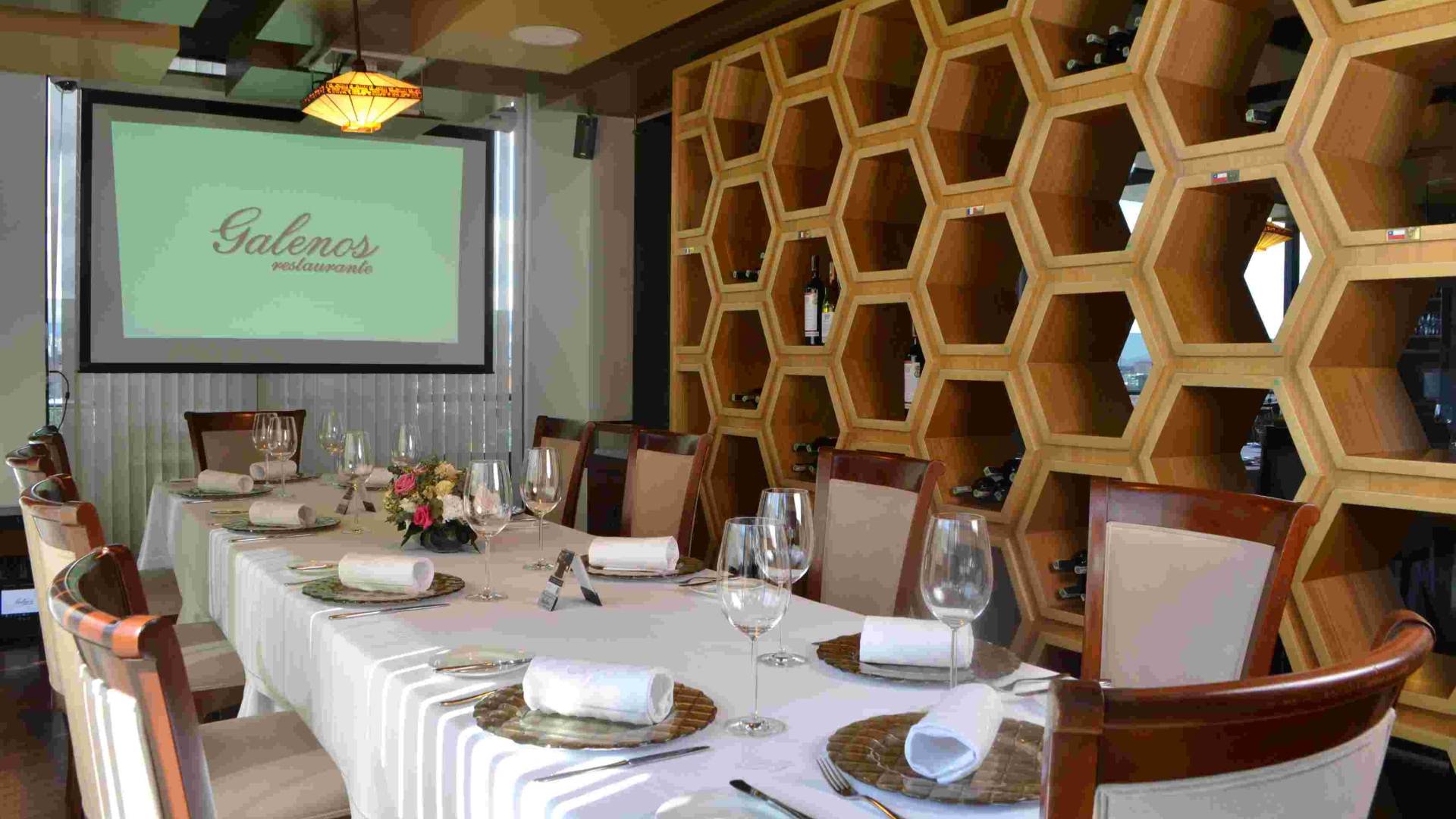 Salon con una mesa alargada cubierta con mantel blanco y con copas de cristal y platos sobre ella. Se ve una cava a un lado y en el fondo una pantalla gigante.