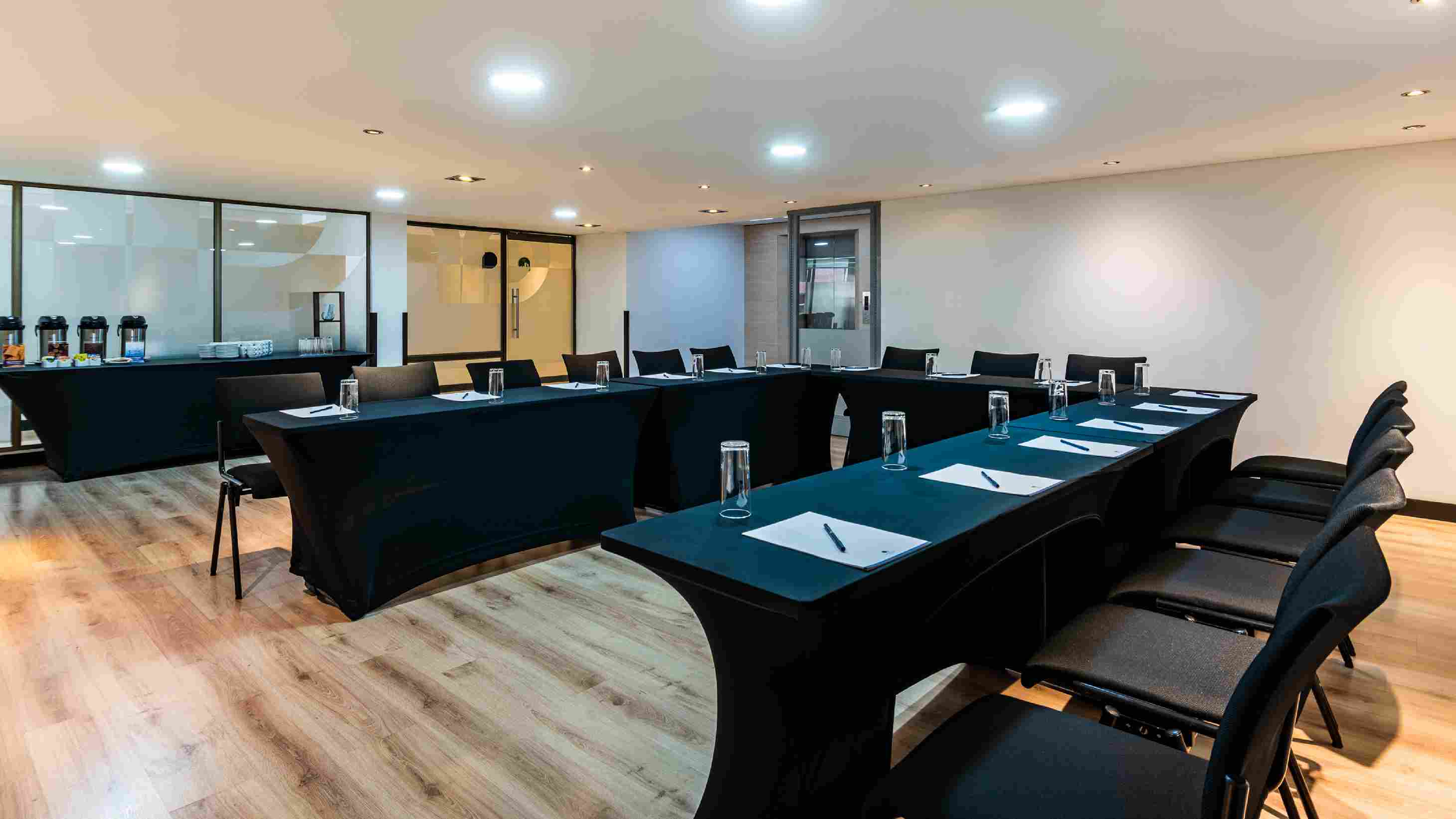Salón con una mesa en U, rodeada por sillas. En la mesa hay vasos de cristal, esferos y papel. A un lado hay mesa con cafeteras.