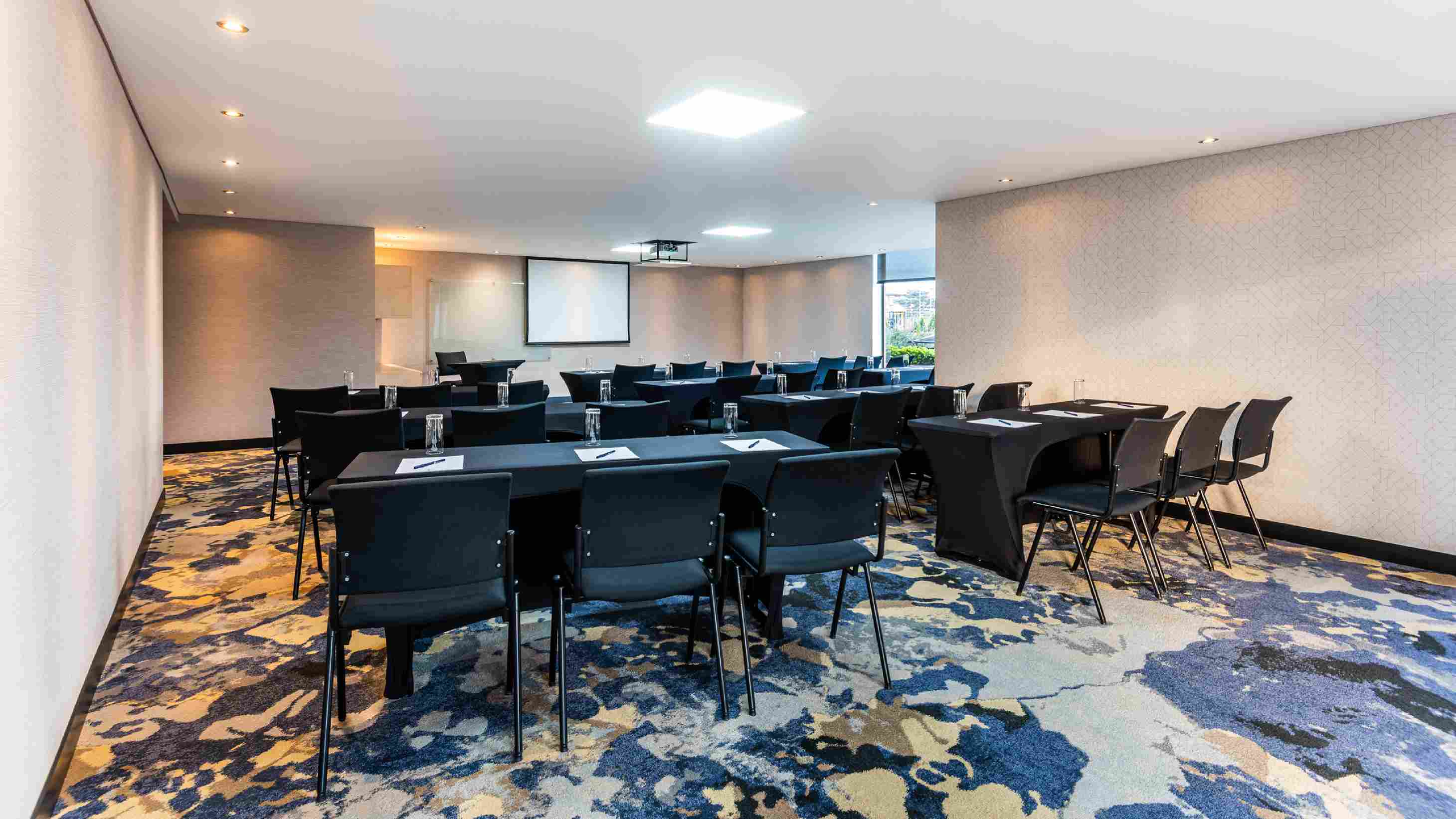  Salón con por lo menos cinco mesas rodeadas por sillas. En las mesas hay vasos de cristal, esferos y papel. Al fondo hay una pantalla de videobeam.