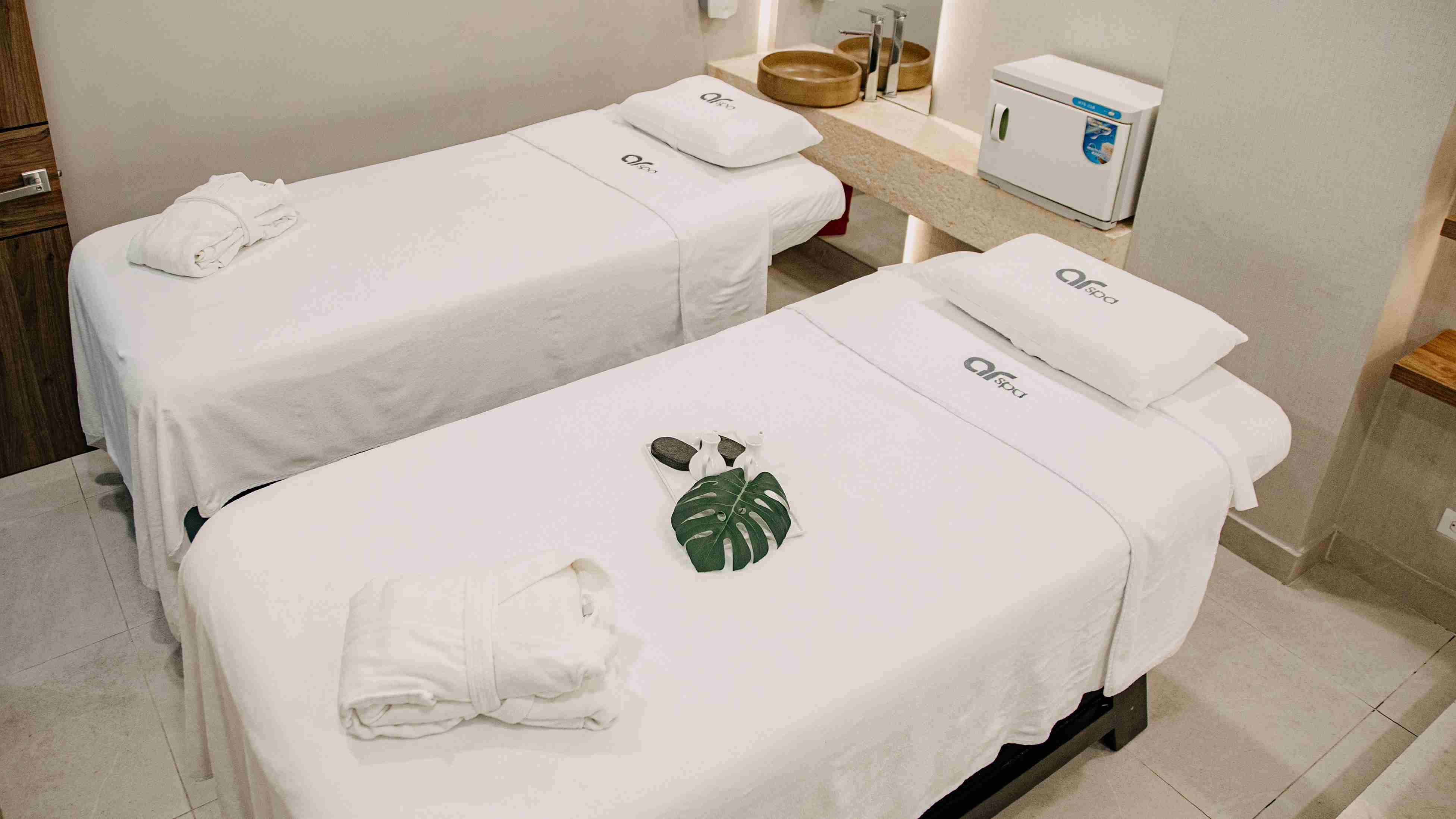 A small room with two massage beds covered with white blankets. Two white robes are placed on each bed. The blankets display the text: “ar spa.”