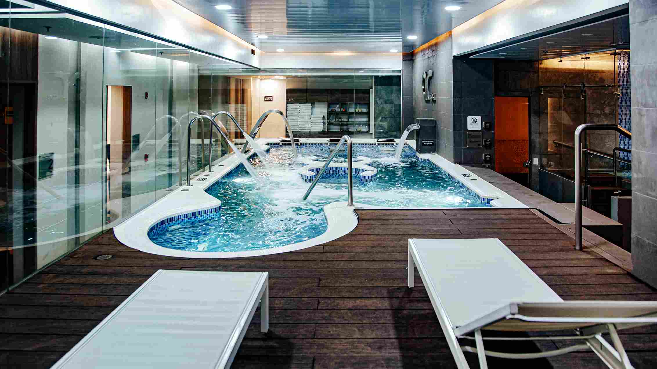 A room with a pool where the water is moving, creating a white layer of bubbles. The space is surrounded by large windows and includes two long reclining lounge chairs.