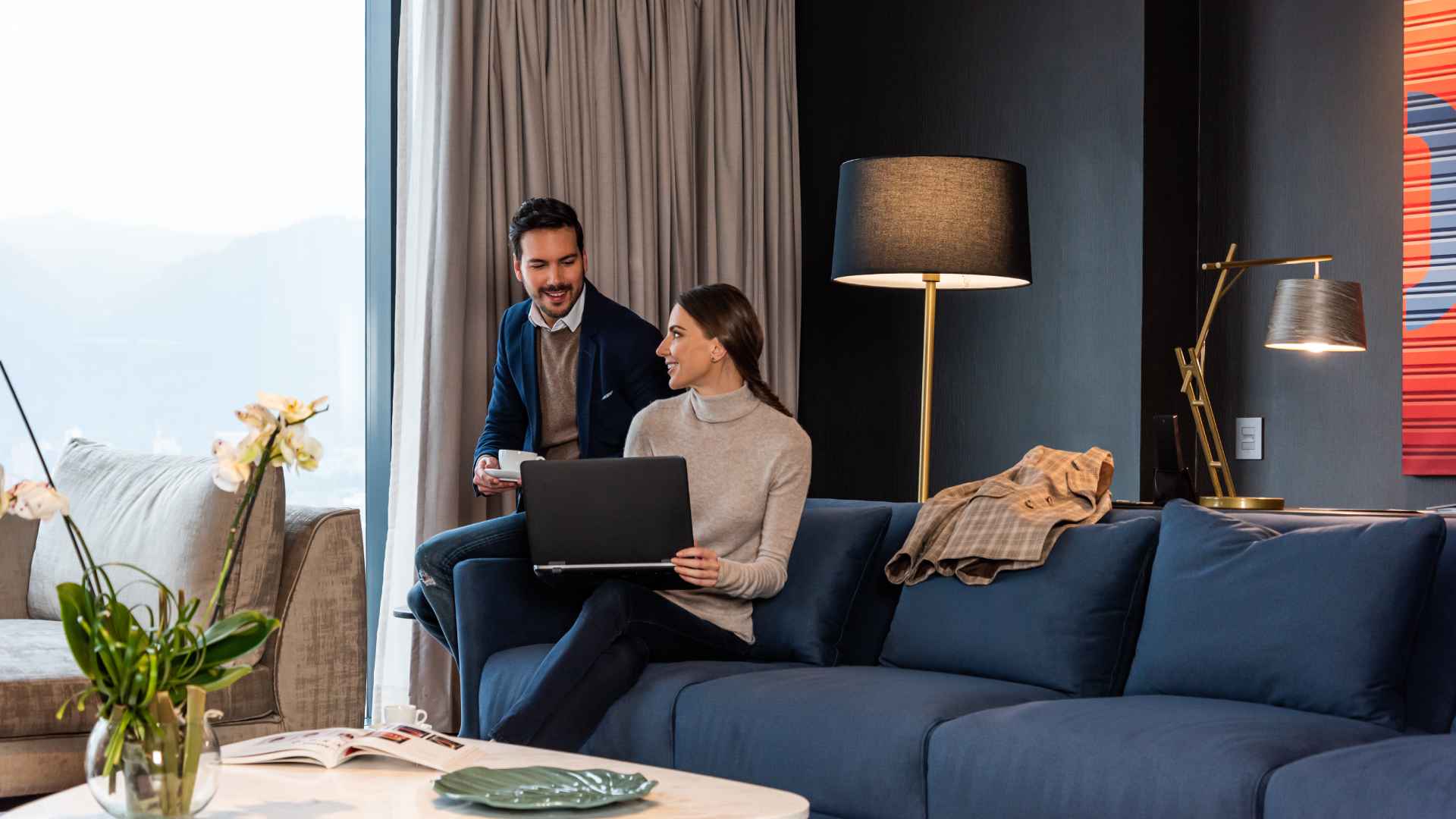  Un hombre y una mujer joven en un salón, sentados en un sofá amplio y observando algo en un computador portátil.