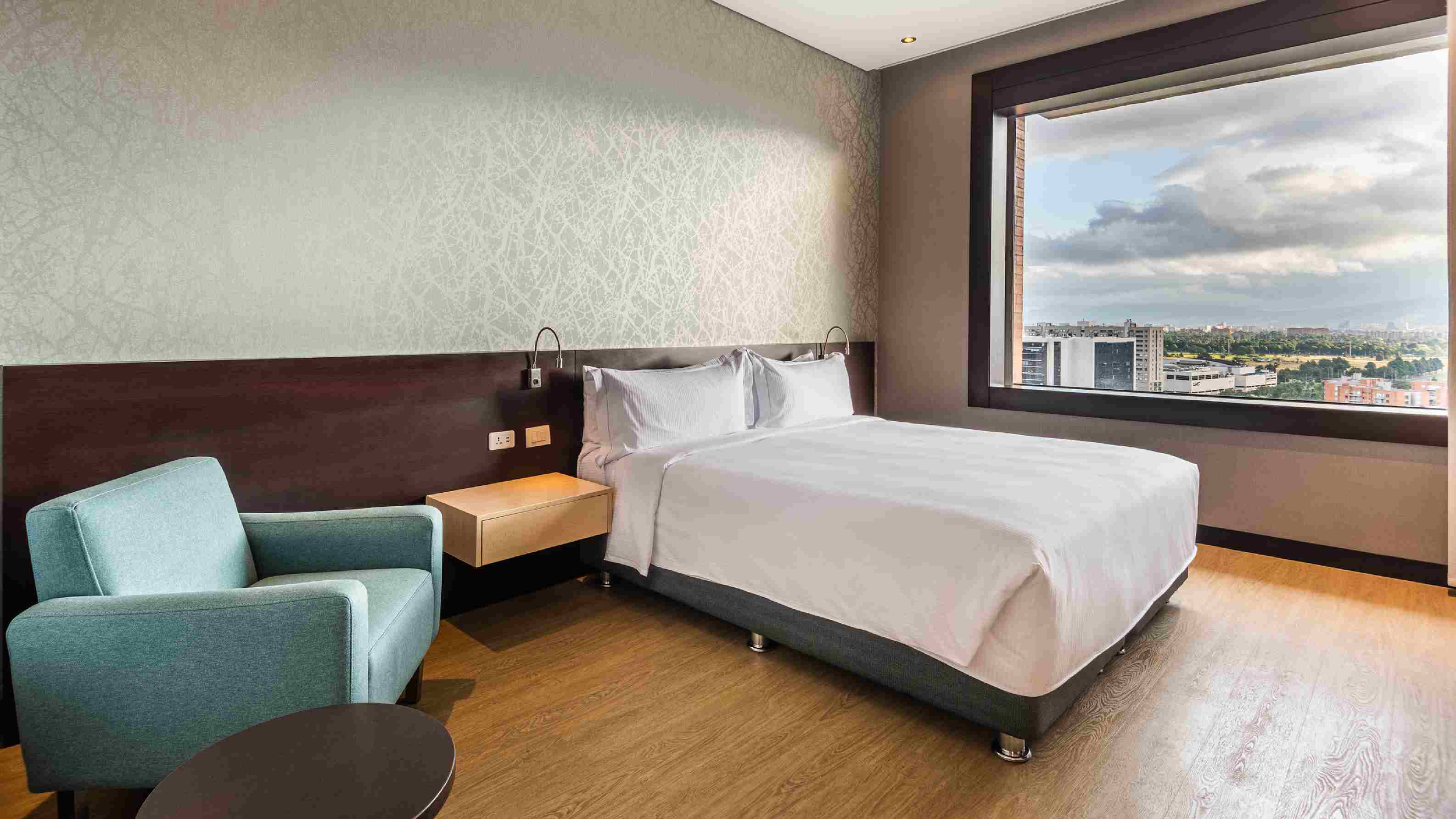  A room on a high floor with a bed dressed in white sheets and a light blue armchair. There is a window without curtains, from which the rooftops of other buildings can be seen in the distance. A room on a high floor with a bed dressed in white sheets and a light blue armchair. There is a window without curtains, from which the rooftops of other buildings can be seen in the distance.