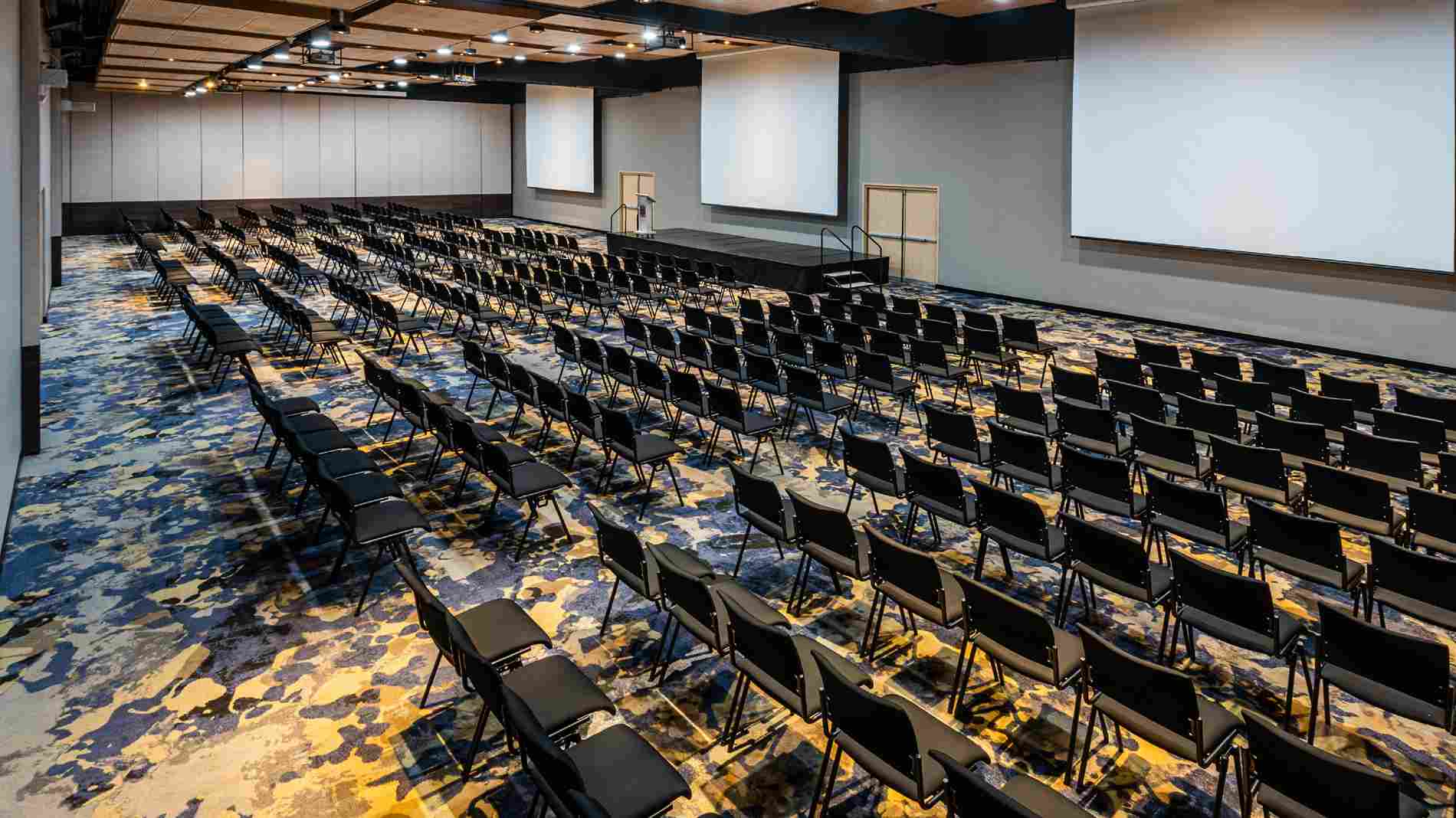  Gran salón con por lo menos un centenar de sillas, todas de frente a tres pantallas gigantes de videobeam. 