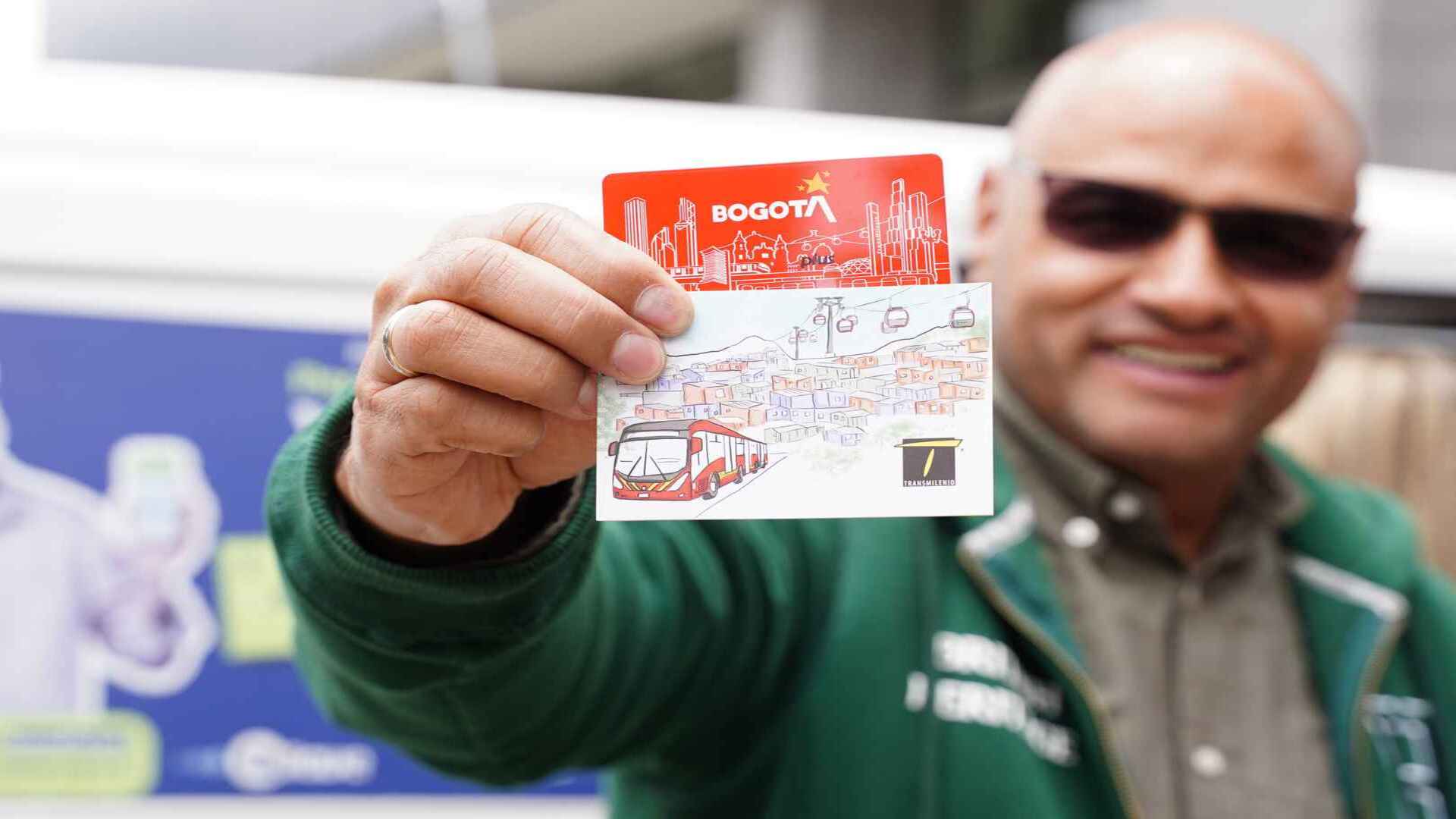 Un hombre sonriente con gafas de sol mostrando una tarjeta de transporte de Bogotá con un diseño de la ciudad. 