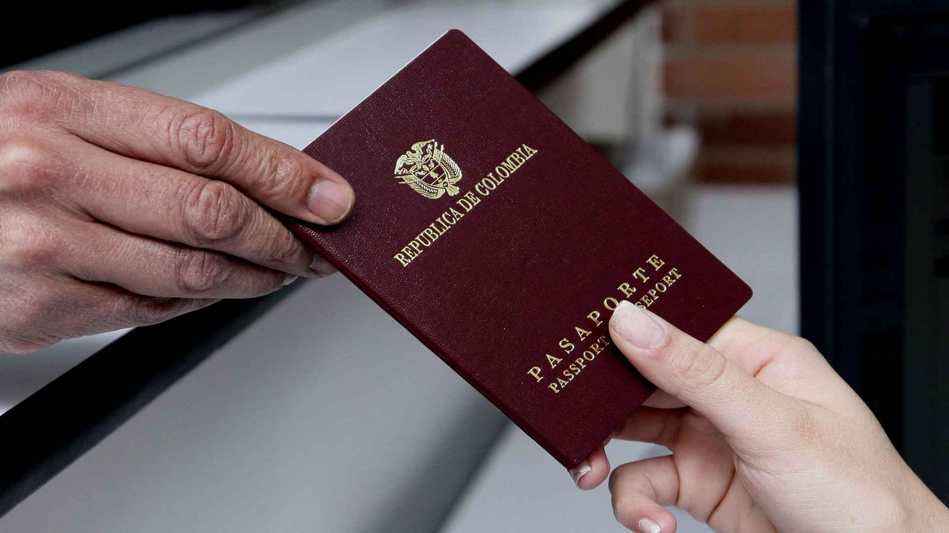 Manos intercambiando un pasaporte colombiano de color vino tinto, simbolizando trámites y viajes.