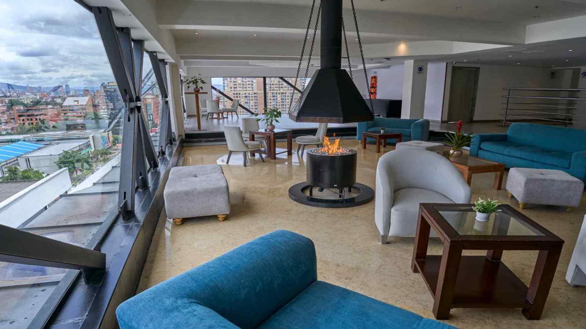 Sala lounge de un hotel con amplios ventanales y vista panorámica de la ciudad, equipada con sofás, mesas y una chimenea central que crea un ambiente moderno, cálido y confortable para el descanso y la conversación.