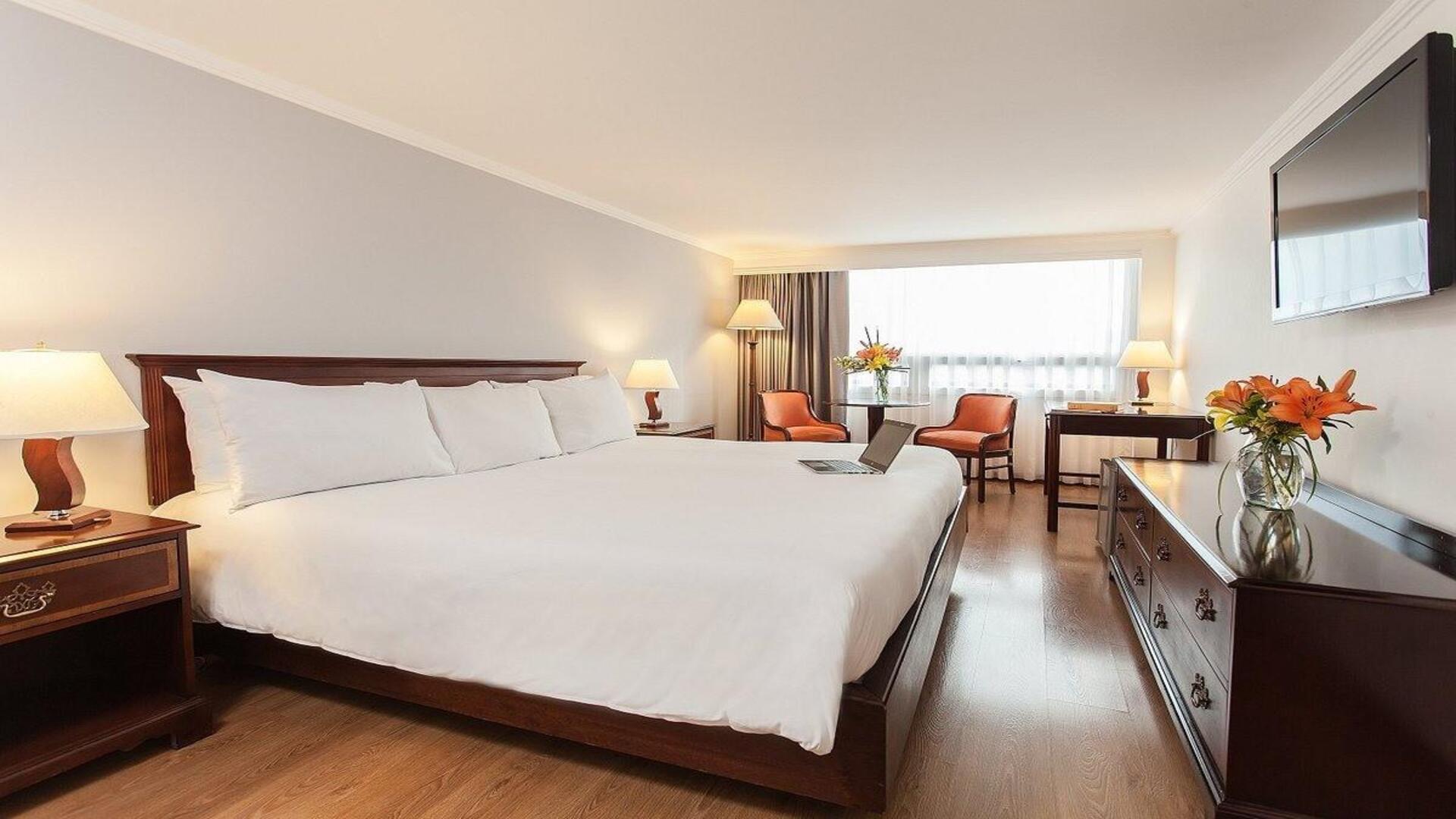 Habitación de hotel amplia y luminosa con cama king size, mobiliario de madera y una decoración elegante que ofrece un ambiente cómodo y acogedor para el descanso.
