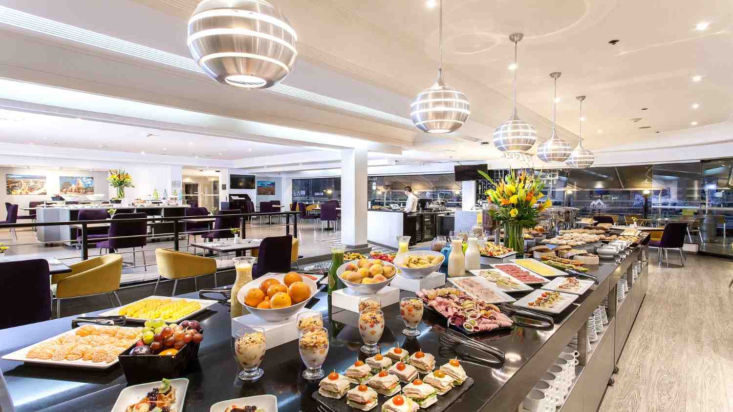 Buffet de restaurante con una amplia variedad de frutas, panes y preparaciones gastronómicas dispuestas en un ambiente moderno y elegante, iluminado con lámparas colgantes y espacios cómodos para los comensales.