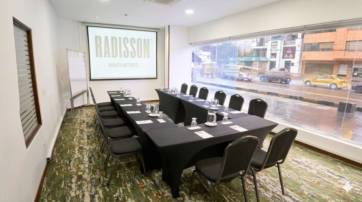 Sala de reuniones moderna con disposición en forma de U, equipada con mesas, sillas y pantalla de proyección, iluminada por amplios ventanales con vista urbana que crean un ambiente profesional y cómodo para encuentros corporativos.