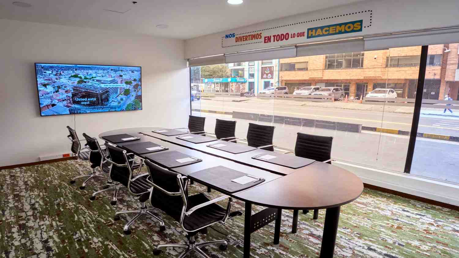 Sala de reuniones moderna con mesa central y sillas ejecutivas, equipada con pantalla digital y amplios ventanales que brindan iluminación natural y vista urbana, en un ambiente cómodo y profesional para encuentros corporativos.