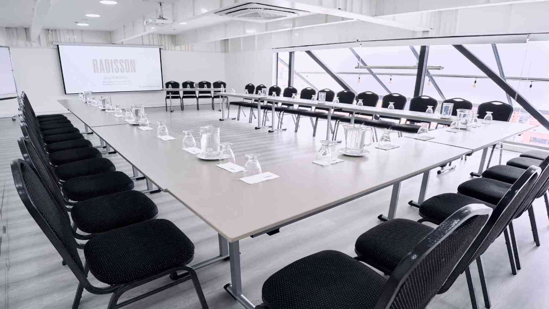  Sala de conferencias amplia y luminosa con disposición de mesas en forma de U, equipada con sillas, pantalla de proyección y ventanales modernos que ofrecen un ambiente profesional ideal para reuniones y eventos corporativos.