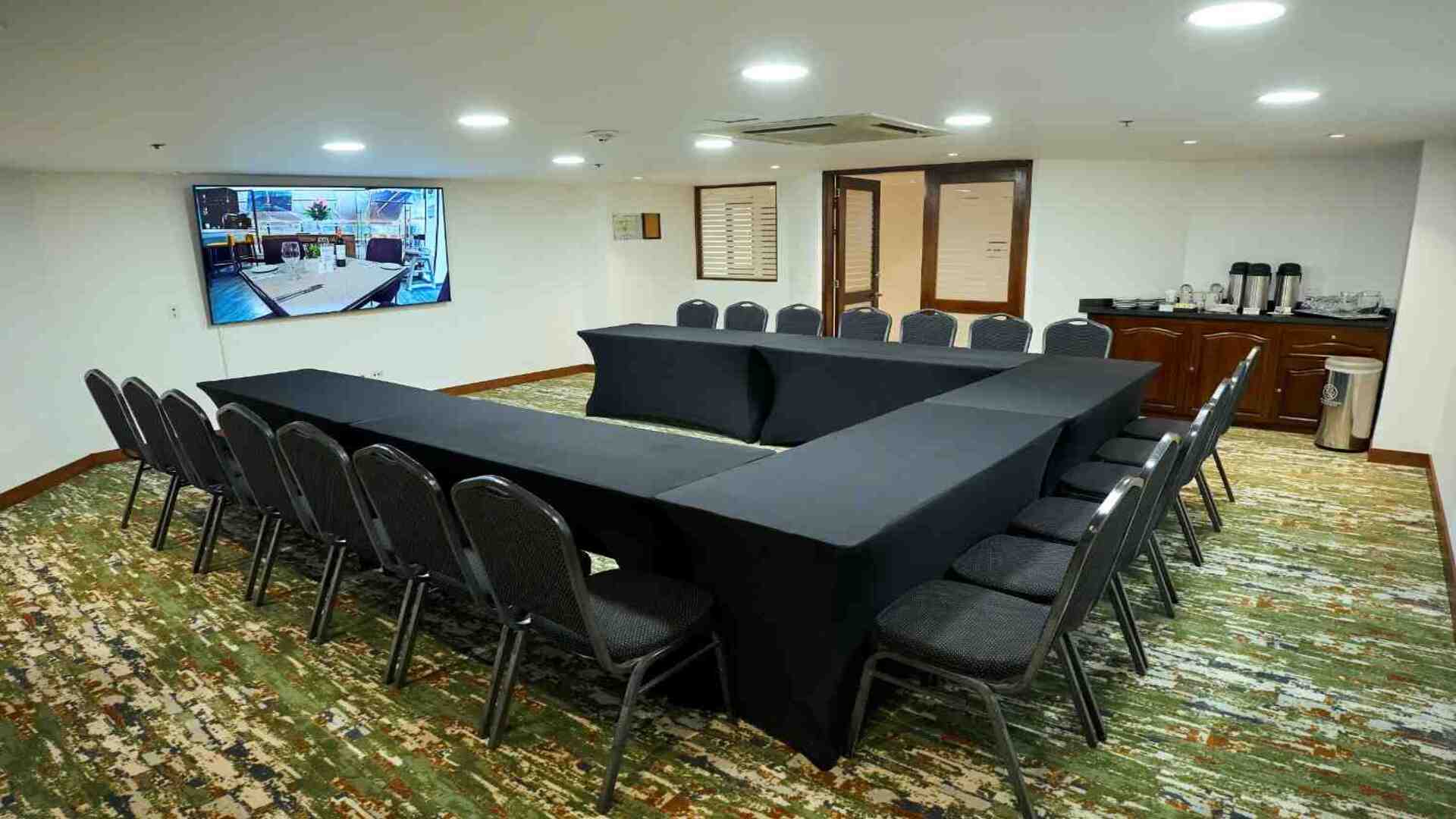 Sala de reuniones con mesas organizadas en forma de U, equipada con pantalla digital, sillas y estación de café, en un ambiente funcional y cómodo para encuentros corporativos y capacitaciones.