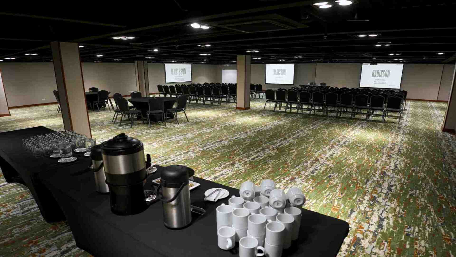 Salón amplio para eventos y conferencias, equipado con filas de sillas, pantallas de proyección y estación de café, diseñado para reuniones corporativas, capacitaciones y encuentros empresariales en un ambiente moderno y funcional.