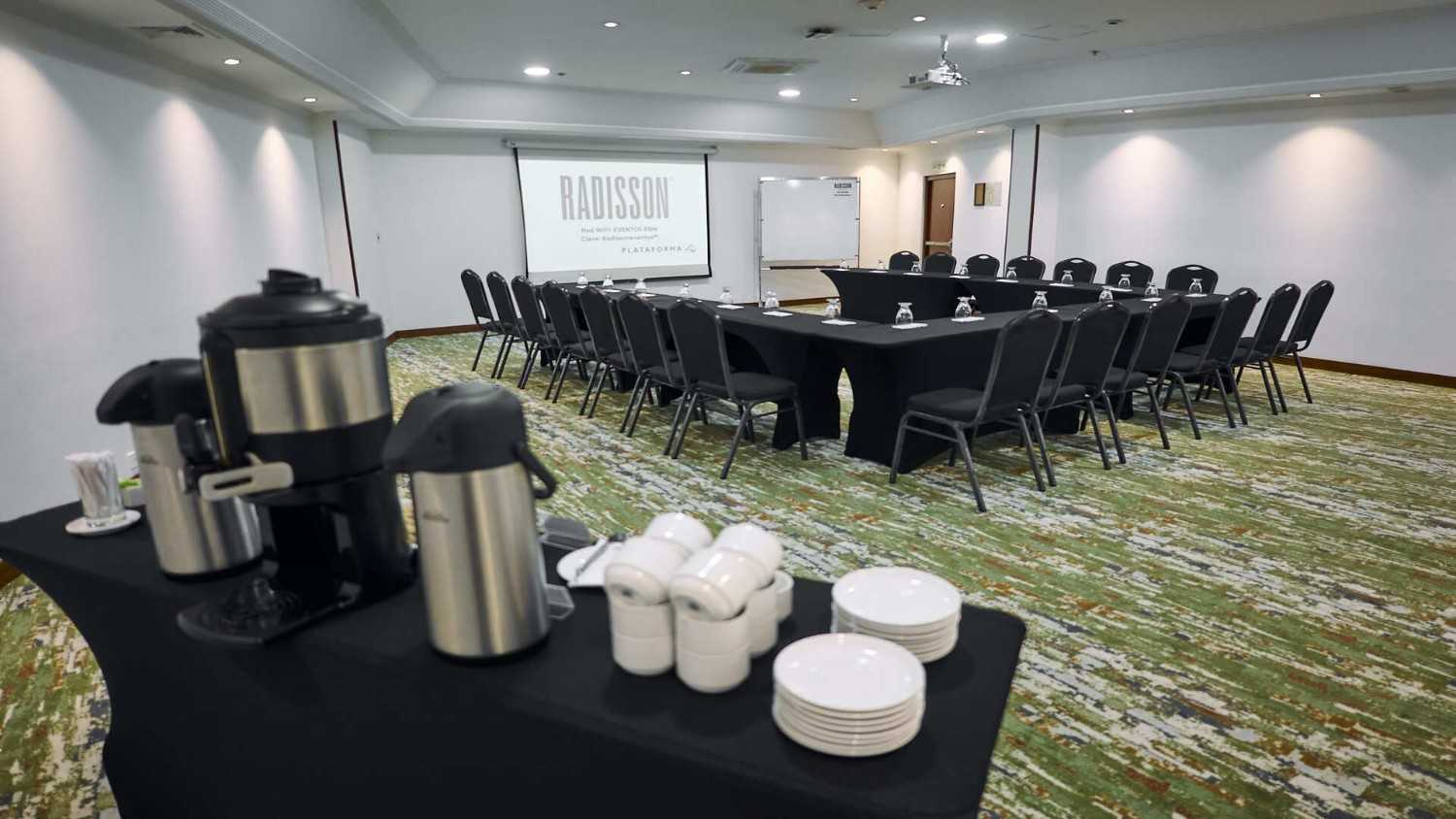 Sala de reuniones corporativa con mesas organizadas en forma de U, equipada con pantalla de proyección, sillas y estación de café, en un espacio amplio y funcional ideal para capacitaciones y encuentros empresariales.