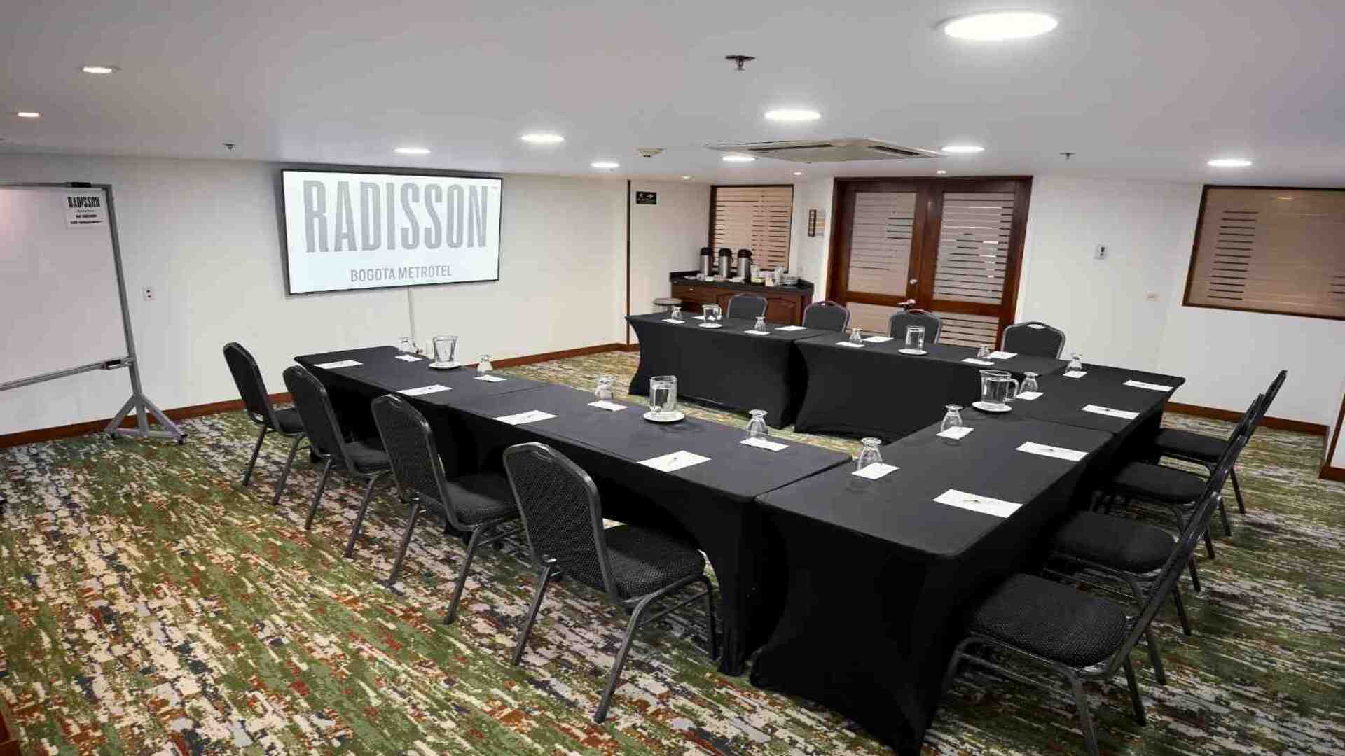  Sala de reuniones ejecutiva con mesas organizadas en forma de U, equipada con pantalla de proyección, tablero y servicio de café, en un ambiente moderno y adecuado para encuentros corporativos y capacitaciones.
