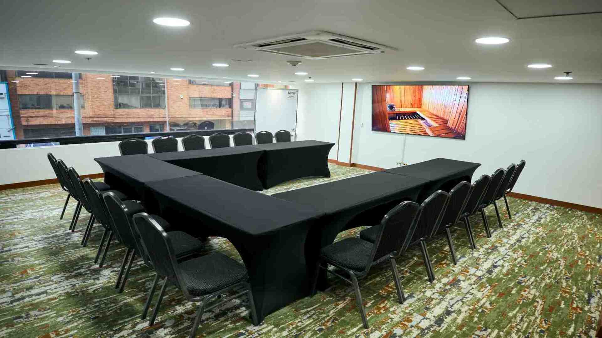  Sala de reuniones moderna con mesas dispuestas en forma de U, equipada con pantalla digital, sillas y amplios ventanales que brindan iluminación natural, ideal para encuentros corporativos y capacitaciones.