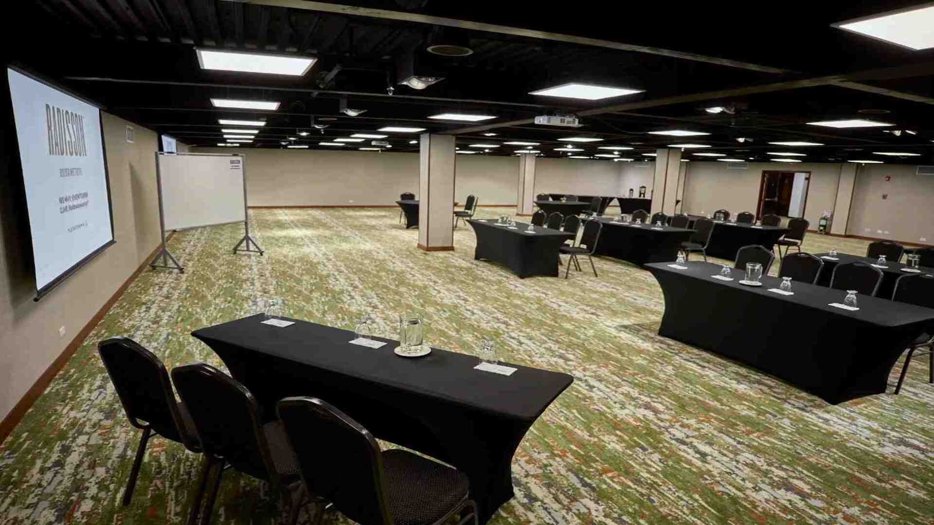 Salón amplio para eventos y reuniones corporativas, con mesas distribuidas en diferentes formatos, pantalla de proyección y espacios abiertos que brindan un ambiente moderno y funcional para conferencias y capacitaciones.