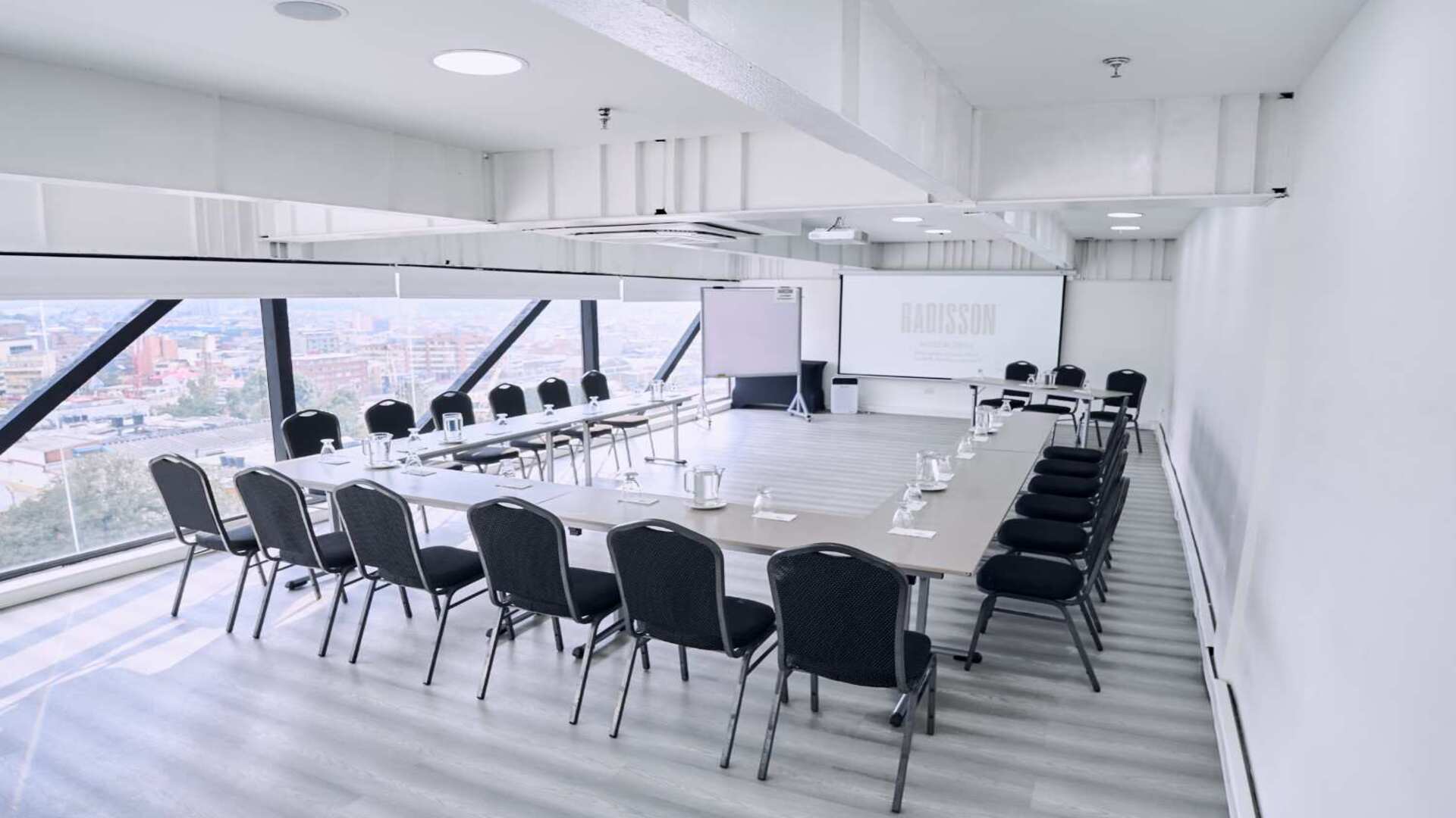 Sala de reuniones elegante y luminosa con mesas organizadas en forma de U, amplios ventanales con vista panorámica de la ciudad y equipamiento para presentaciones, ideal para encuentros corporativos y capacitaciones.