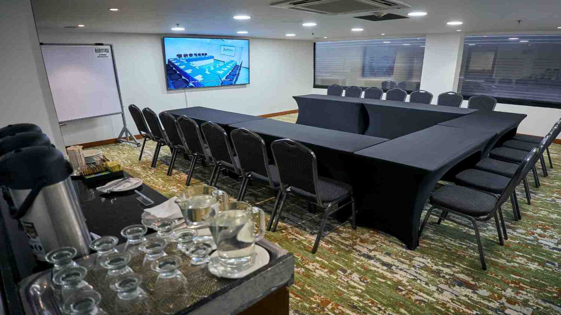 Sala de reuniones moderna con mesas organizadas en forma de U, equipada con pantalla digital, tablero y estación de bebidas, en un ambiente cómodo y funcional para capacitaciones y encuentros corporativos.
