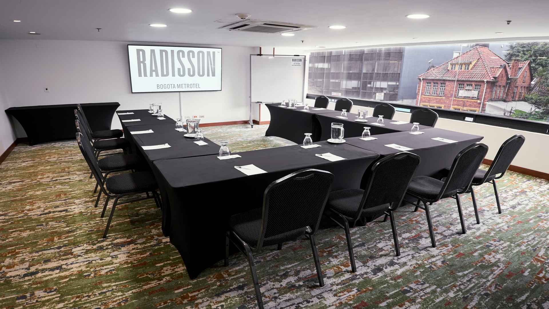 Sala de reuniones ejecutiva con mesas organizadas en forma de U, equipada con pantalla de proyección, tablero y amplios ventanales con vista urbana, ideal para encuentros corporativos y capacitaciones en un ambiente moderno y profesional.