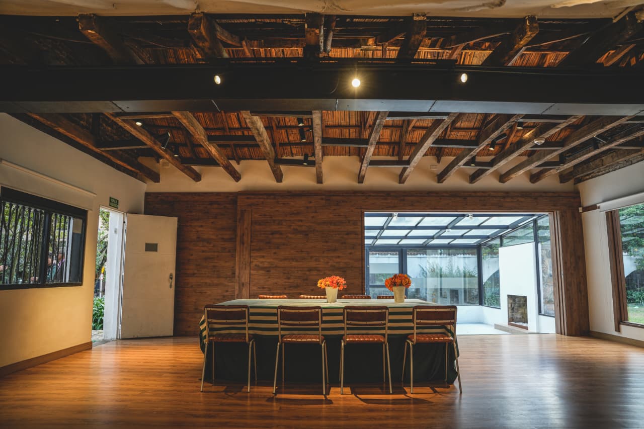 Un amplio salón de estilo colonial combina pisos de madera, vigas expuestas y abundante luz natural. La mesa central, decorada con arreglos florales, resalta un espacio elegante y versátil, ideal para reuniones, eventos culturales y encuentros corporativos.