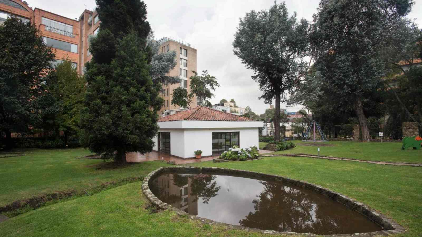 Un amplio jardín rodea una construcción de estilo colonial, junto a un pequeño estanque que refleja el paisaje. Los árboles centenarios y las zonas verdes crean un ambiente tranquilo que invita al descanso y al contacto con la naturaleza en medio de la ciudad.