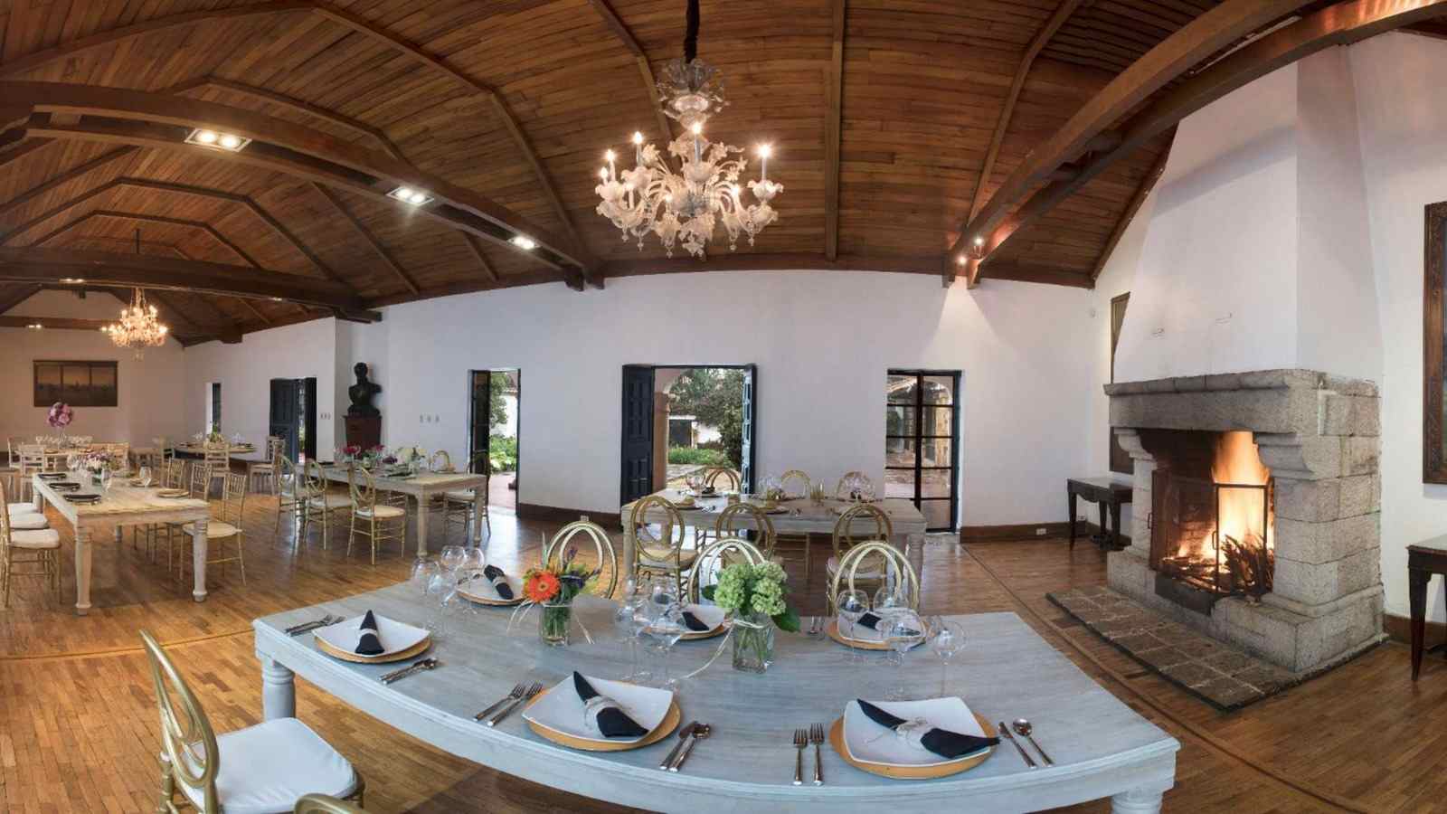 Un amplio salón de estilo colonial combina techos de madera, lámparas de cristal y una acogedora chimenea de piedra. Las mesas elegantemente dispuestas crean un ambiente cálido y sofisticado, ideal para eventos sociales, culturales y corporativos.