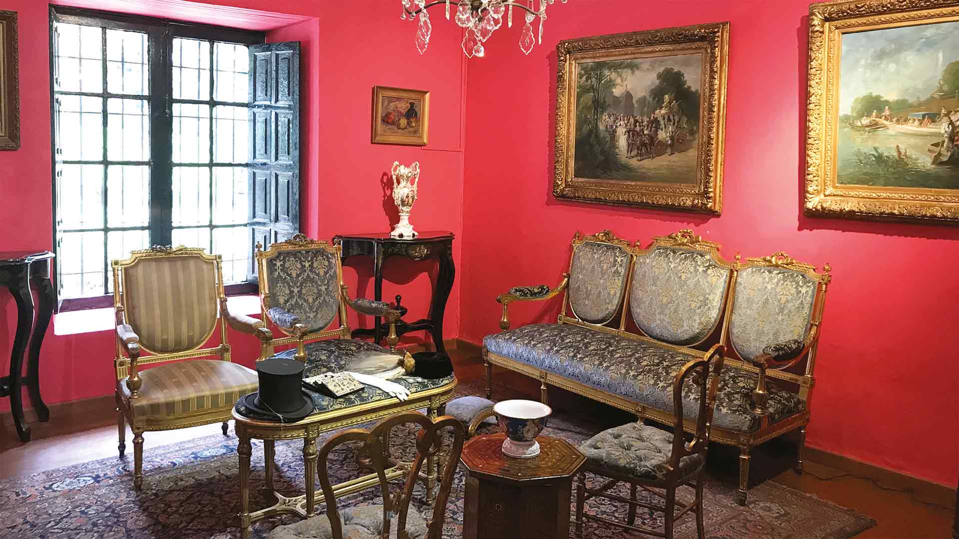 Una elegante sala del Museo El Chicó exhibe mobiliario de época, pinturas enmarcadas y objetos decorativos sobre paredes de intenso color rojo. La luz natural que entra por el ventanal resalta el ambiente histórico y el valor patrimonial de este espacio cultural de Bogotá.
