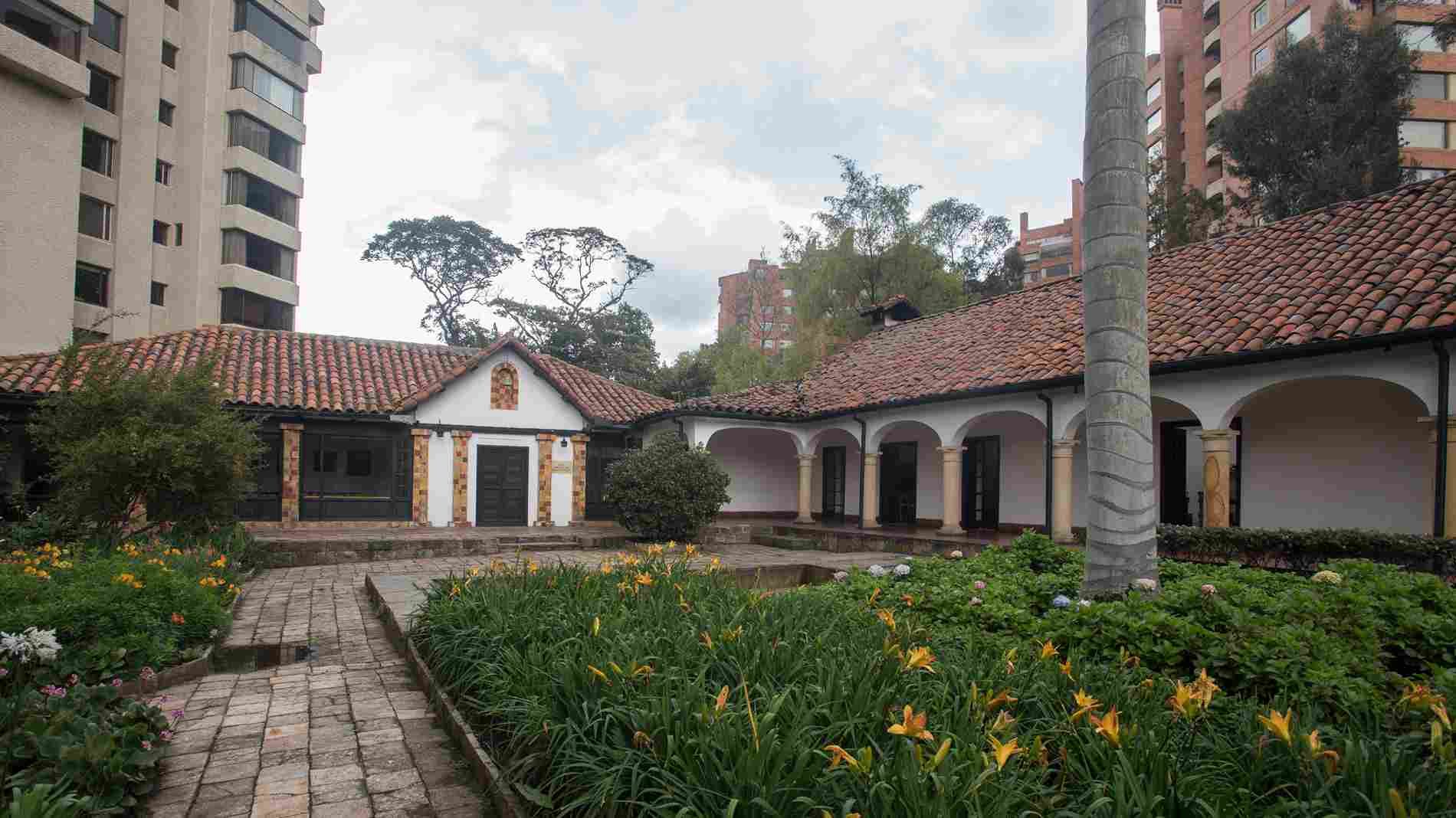 Un patio colonial rodeado de jardines y senderos empedrados enmarca una casona de fachadas blancas, techos de teja y arcos tradicionales. Este espacio histórico ofrece un ambiente sereno donde la arquitectura patrimonial y la naturaleza conviven en pleno corazón de Bogotá.