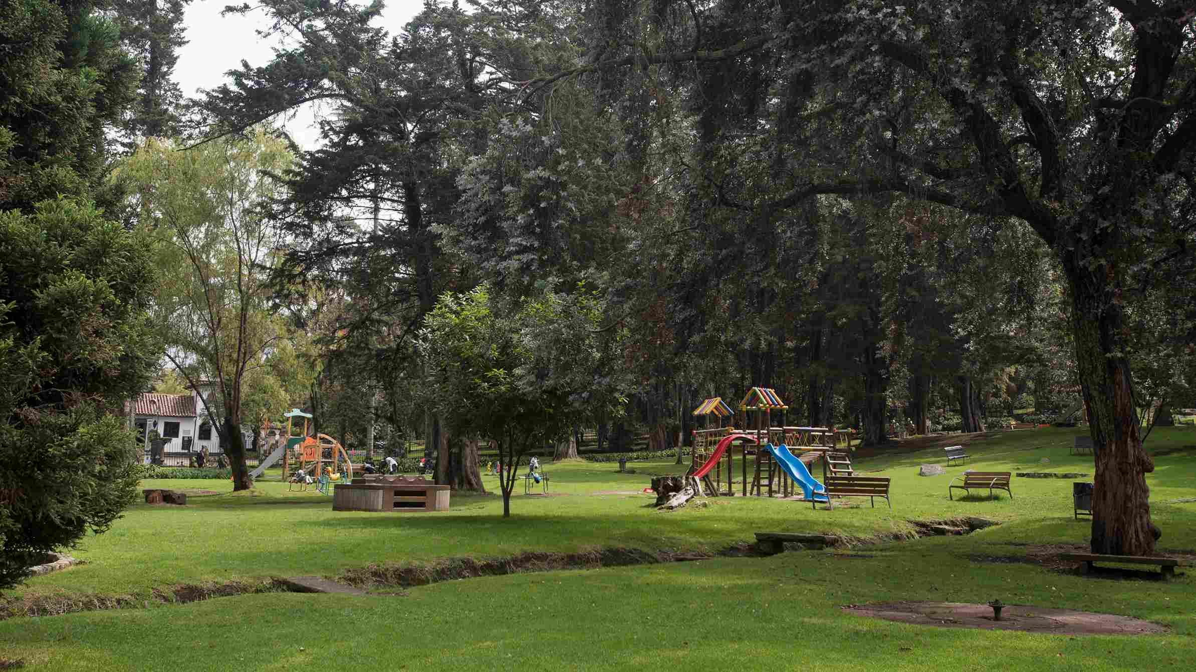 Parque con amplias zonas verdes, árboles de gran tamaño, juegos infantiles de colores, bancas y un pequeño canal de agua que atraviesa el jardín, en un ambiente tranquilo y natural.
