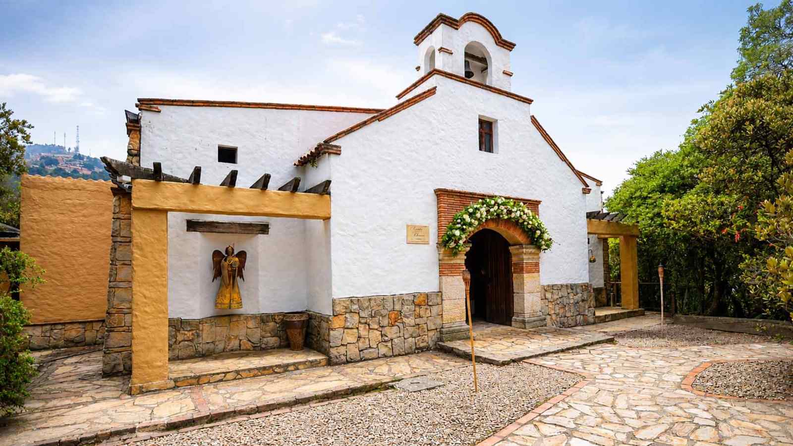 Una encantadora construcción de estilo colonial, con muros blancos, detalles en piedra y ladrillo, y una tradicional torre campanario. El entorno ajardinado y la decoración floral en la entrada resaltan su carácter histórico, acogedor y lleno de encanto.