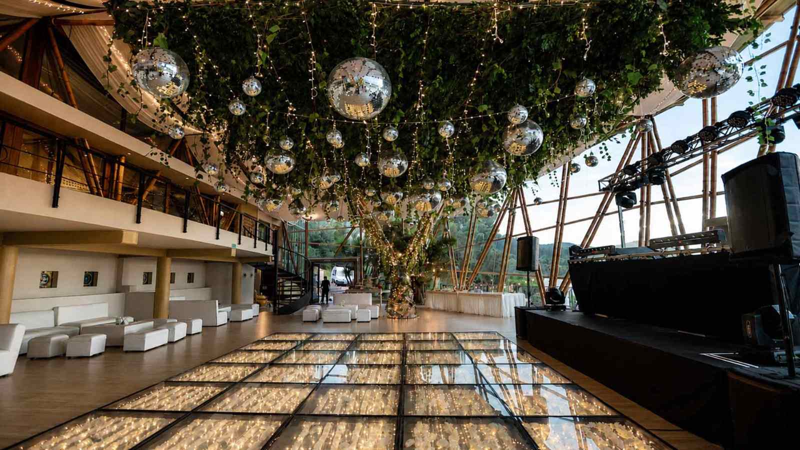 Un escenario elegante y espectacular se prepara para una celebración, con una pista de baile iluminada, cientos de luces y esferas decorativas suspendidas entre la vegetación. La combinación de naturaleza, diseño y tecnología crea una atmósfera sofisticada y memorable para grandes eventos.