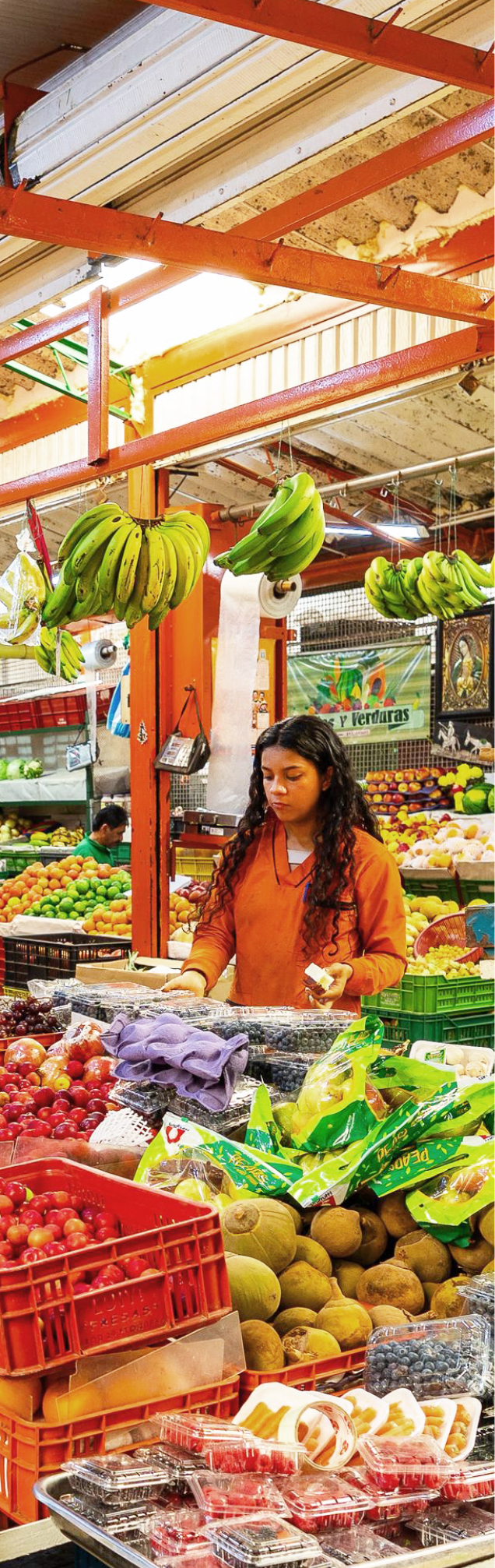 Una mujer joven con una camisa naranja atiende un puesto en un mercado cubierto, rodeada de montones de frutas y verduras frescas y coloridas. Manojos de plátanos verdes cuelgan por encima.