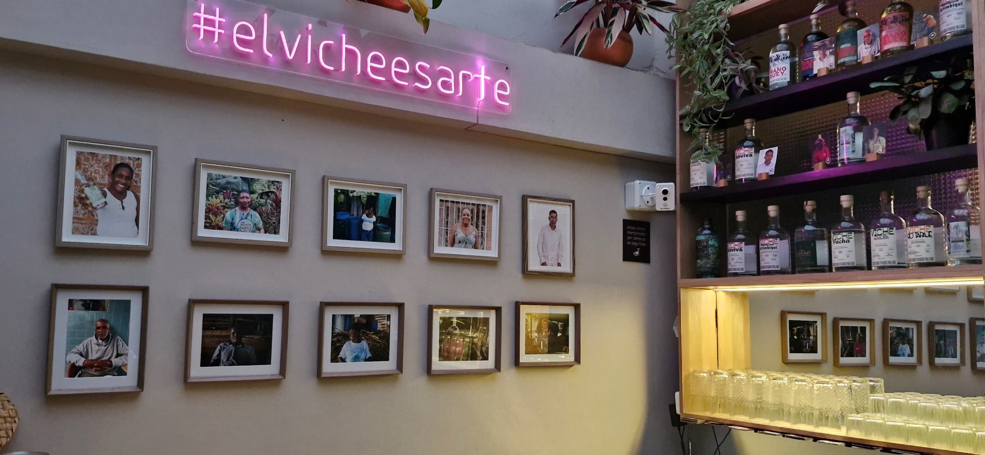 Vichería Pacífico con salón en el que hay una pared con una repisa y varias botellas de licor con viche y varios vasos vacíos. Hay una pared blanca en la que resalta en neón rosa un aviso que dice #elvicheesarte y que está acompañado de varias fotografías de personas de raza negra.