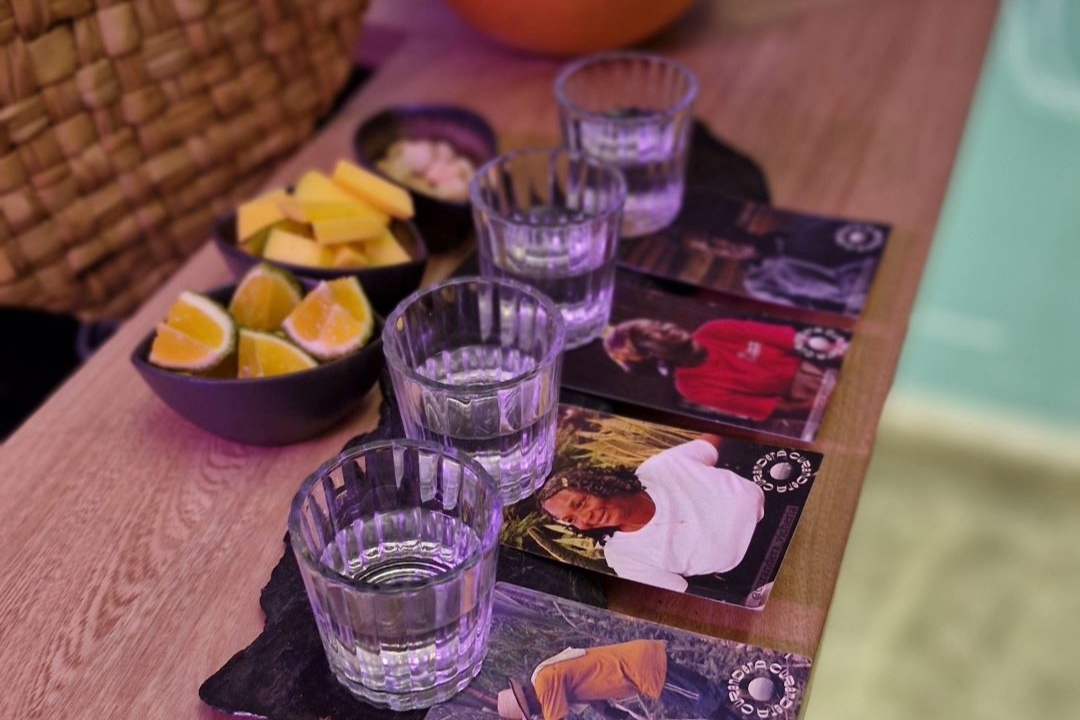 Vichería Pacífico con una mesa con cuatro copas de cristal pequeñas en las que está servido licor, viche. debajo de cada copa hay sendos portavasos con rostros de personas de raza negra. En la mesa hay dos vasijas, una con pedaazos de limón y otra con maní.