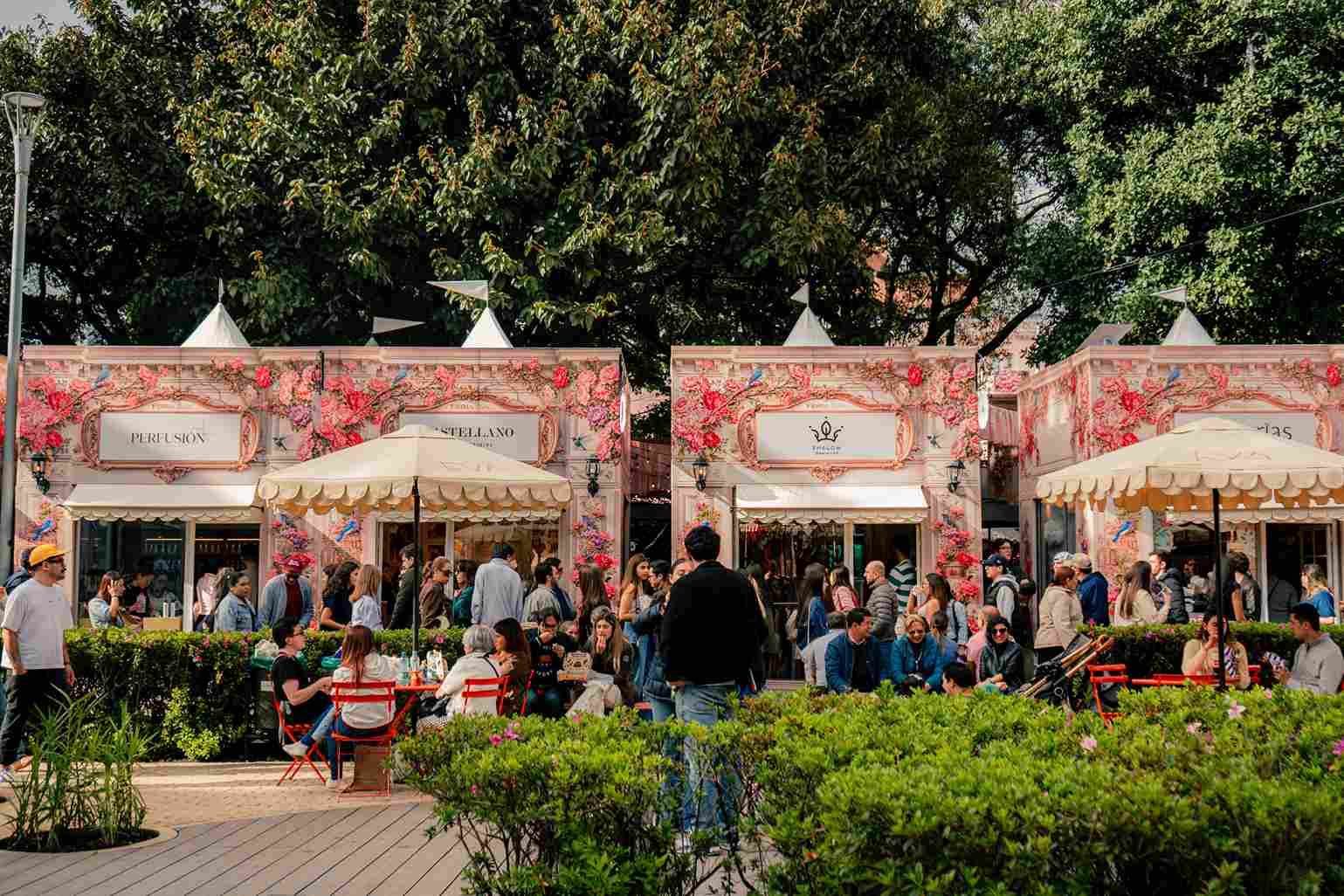 Visitantes recorren una feria al aire libre con coloridos pabellones, restaurantes y zonas de descanso rodeadas de vegetación.