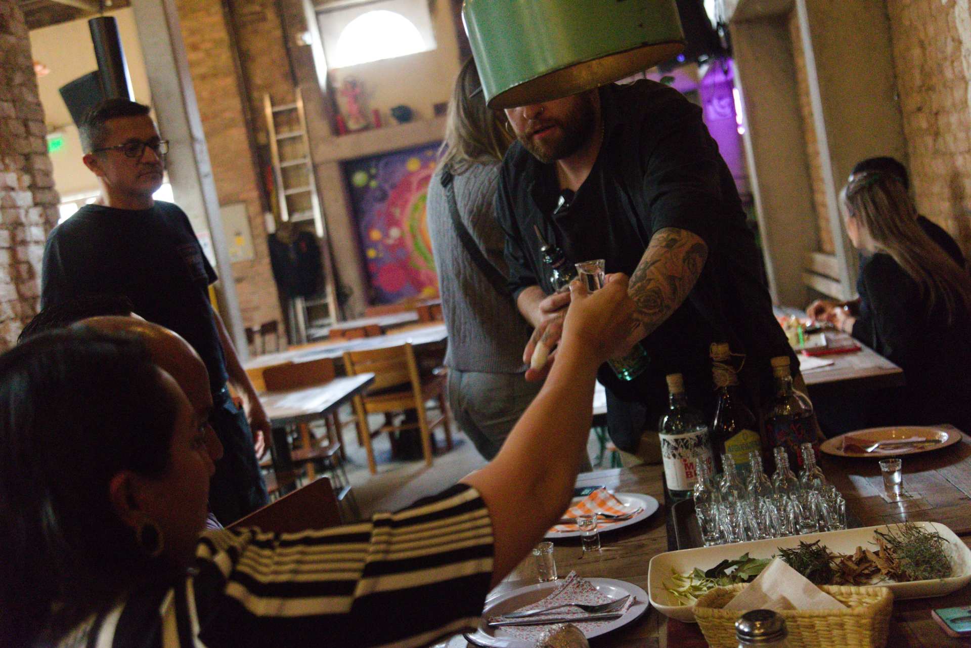 Un hombre joven sentado con una copa de licor con su mano alzada brinda con otro hombre joven. Al fondo hay otras personas que observan. Hay una mesa con varias copas de licor junto con 3 botellas también de licor.
