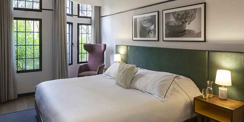 Hotel HAB interior habitación de paredes blancas con cuadros y una cama queen  con tendidos blancos y dos mesas de noche con pantallas encendidas. Una sostiene una botella de agua natural y un vaso. Al lado de la cama una poltrona violeta y al fondo seis ventanas grandes con cortinas abiertas.